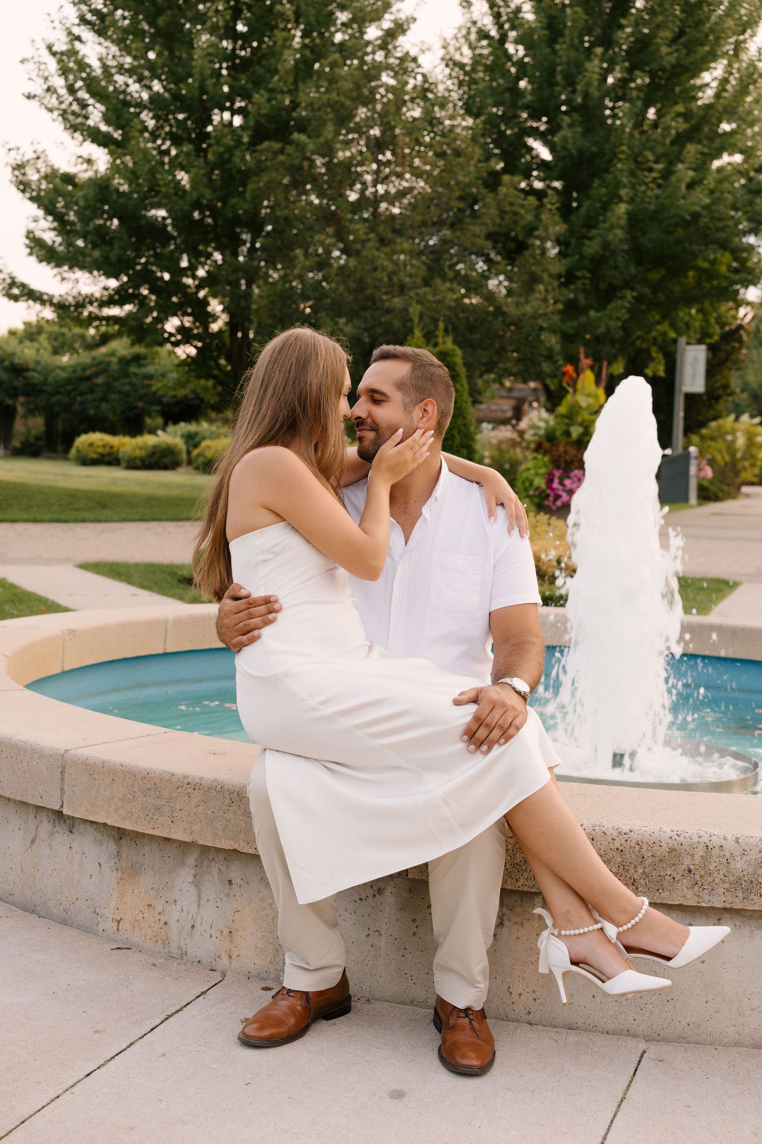 Romantic couple sharing a kiss by a fountain in a park, woman sitting on mans lap at sunset.