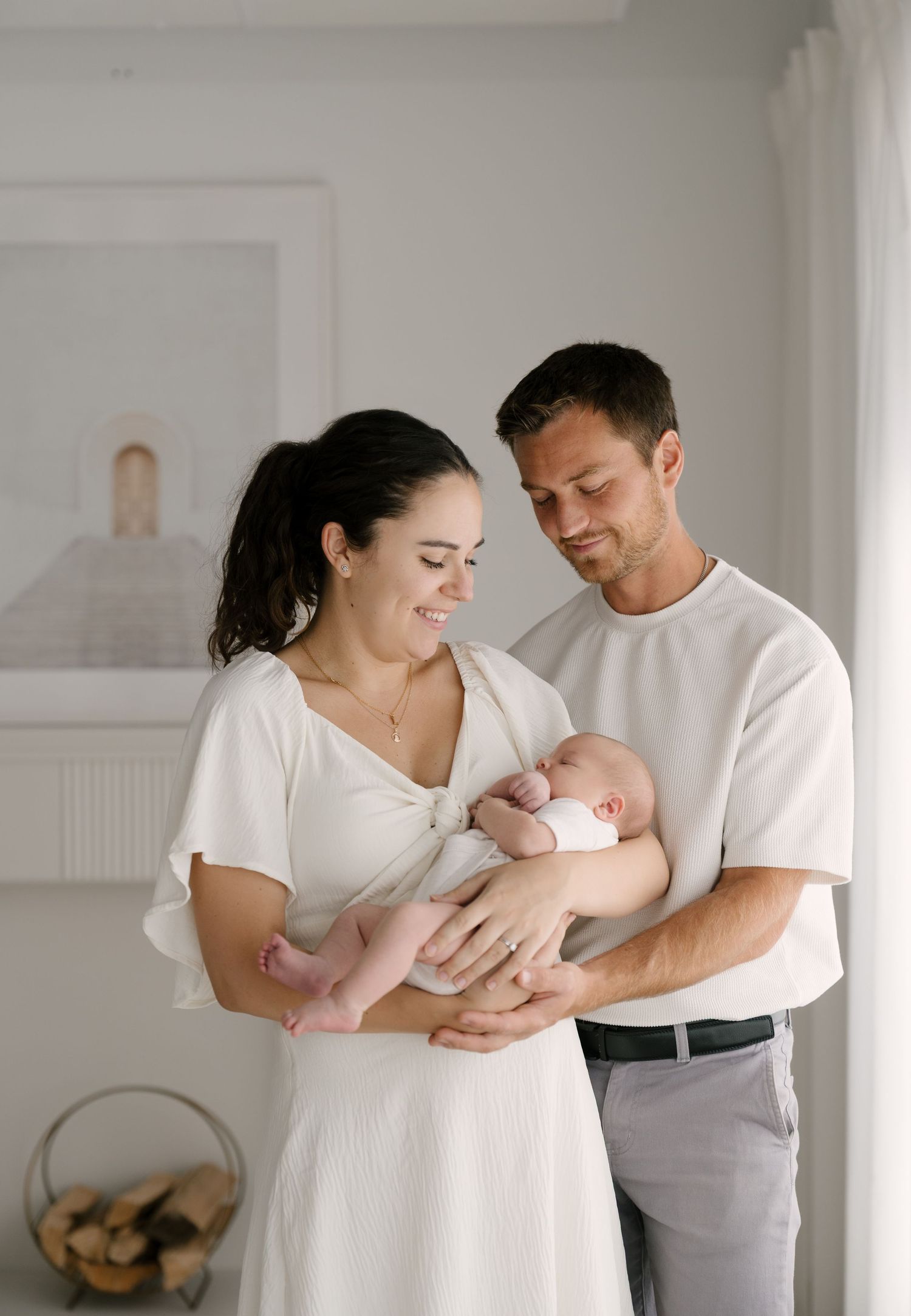 Smiling parents in white outfits tenderly holding their newborn baby indoors in a bright room.