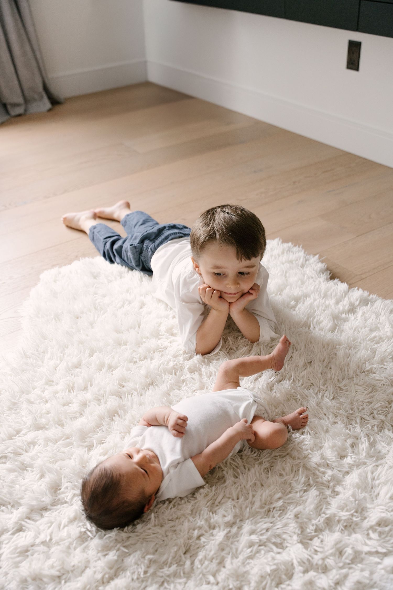 Young boy lying on white fluffy rug smiling at newborn sibling in white onesie beside him.