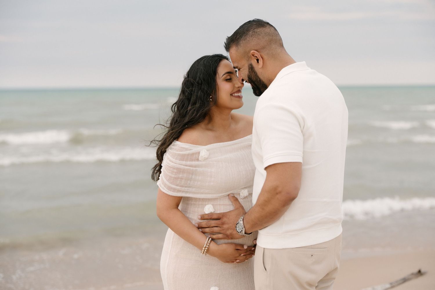 Couple embraces tenderly on beach, pregnant woman in white off-shoulder dress leaning into partners arms.
