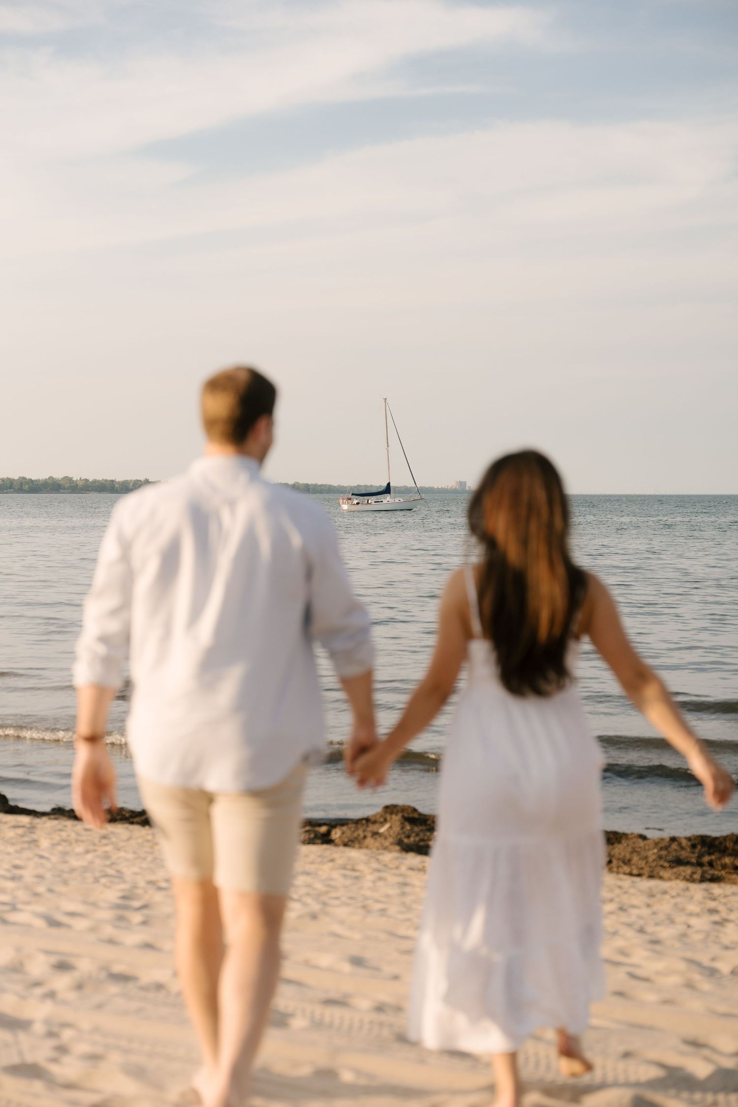 Couple holding hands walking toward ocean on sandy beach with sailboat visible on the horizon.