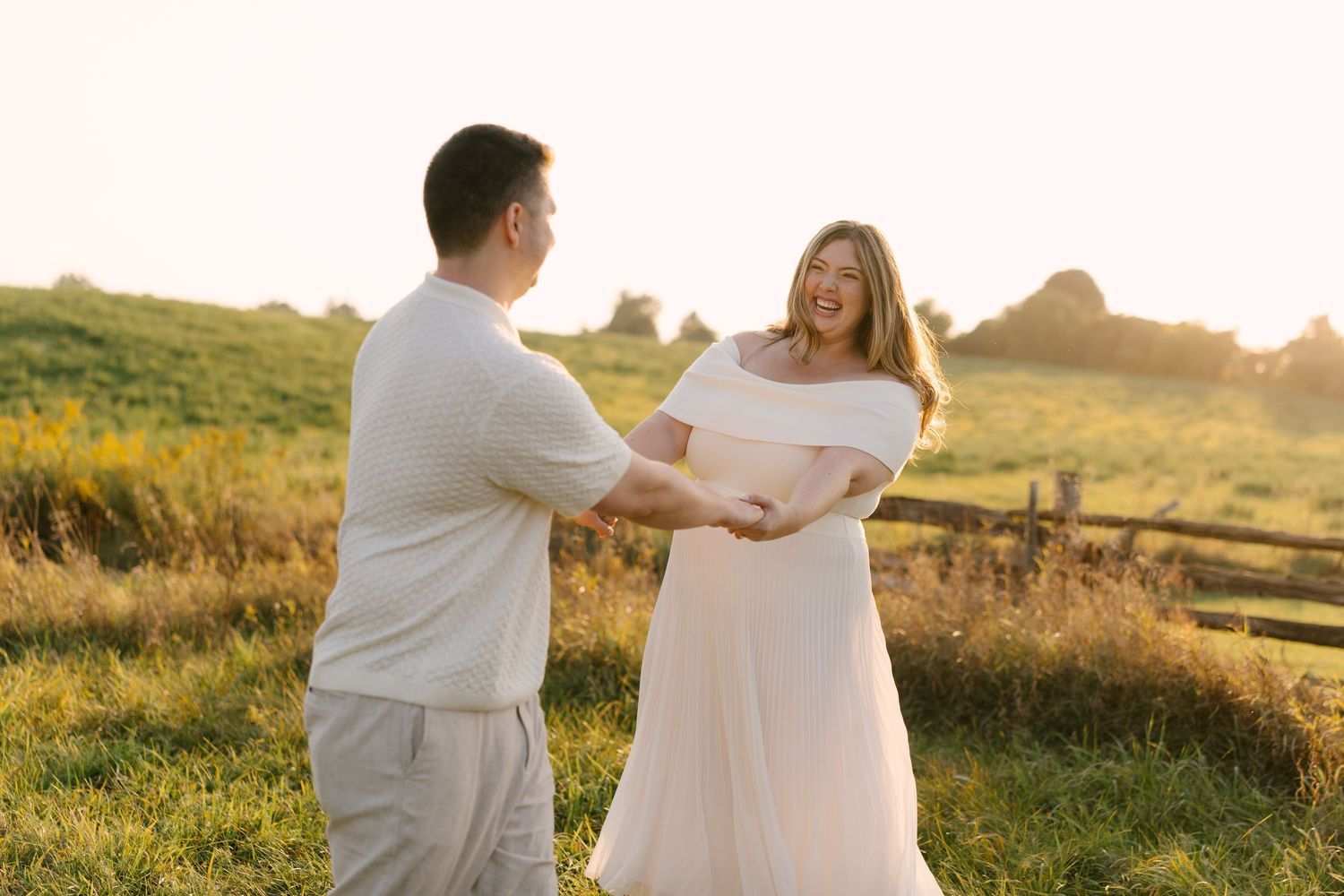 Couple holding hands and laughing together in sunlit grassy field at golden hour.