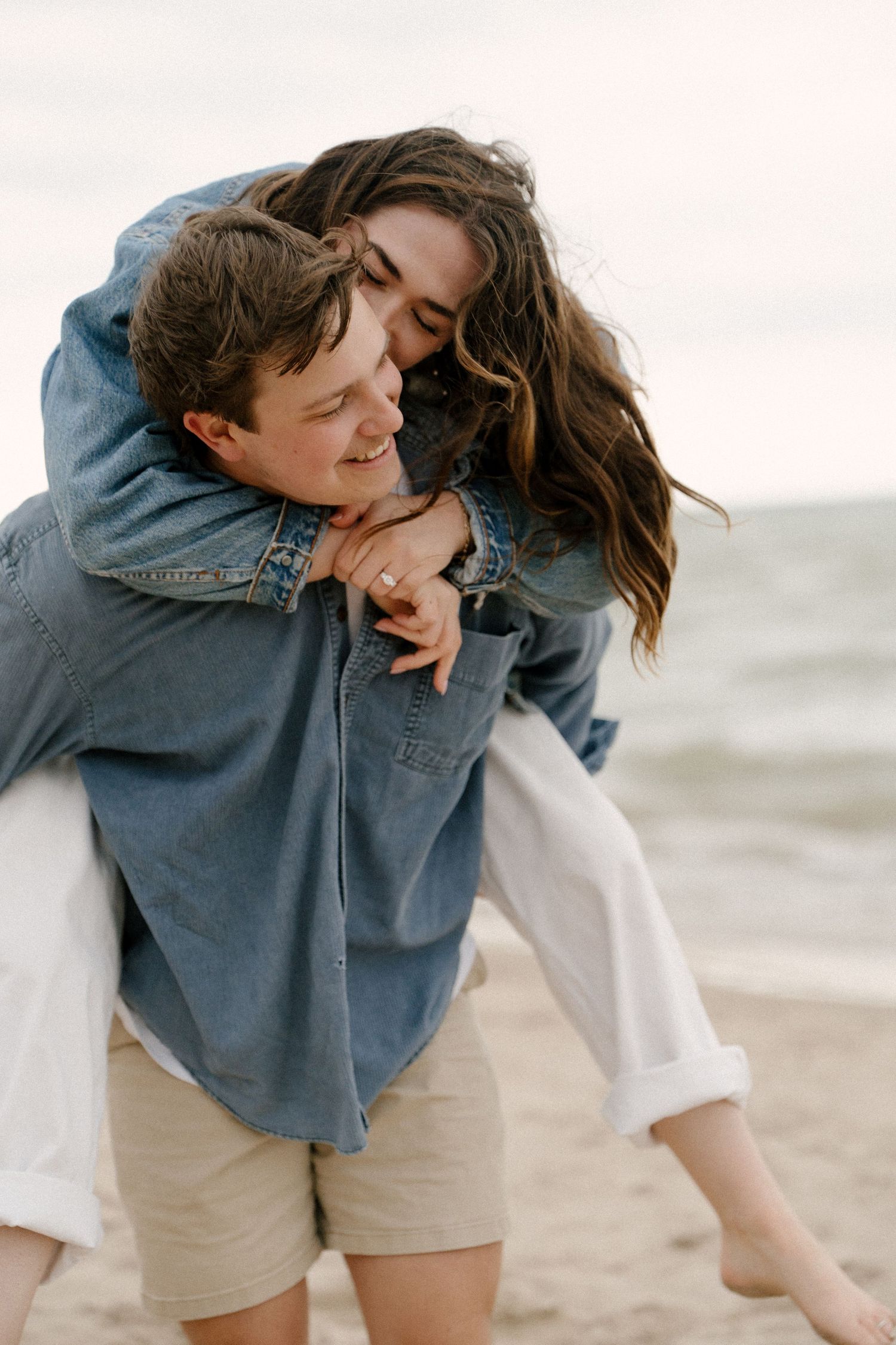 Woman joyfully hugging man from behind in a piggyback embrace on a sandy beach shoreline.