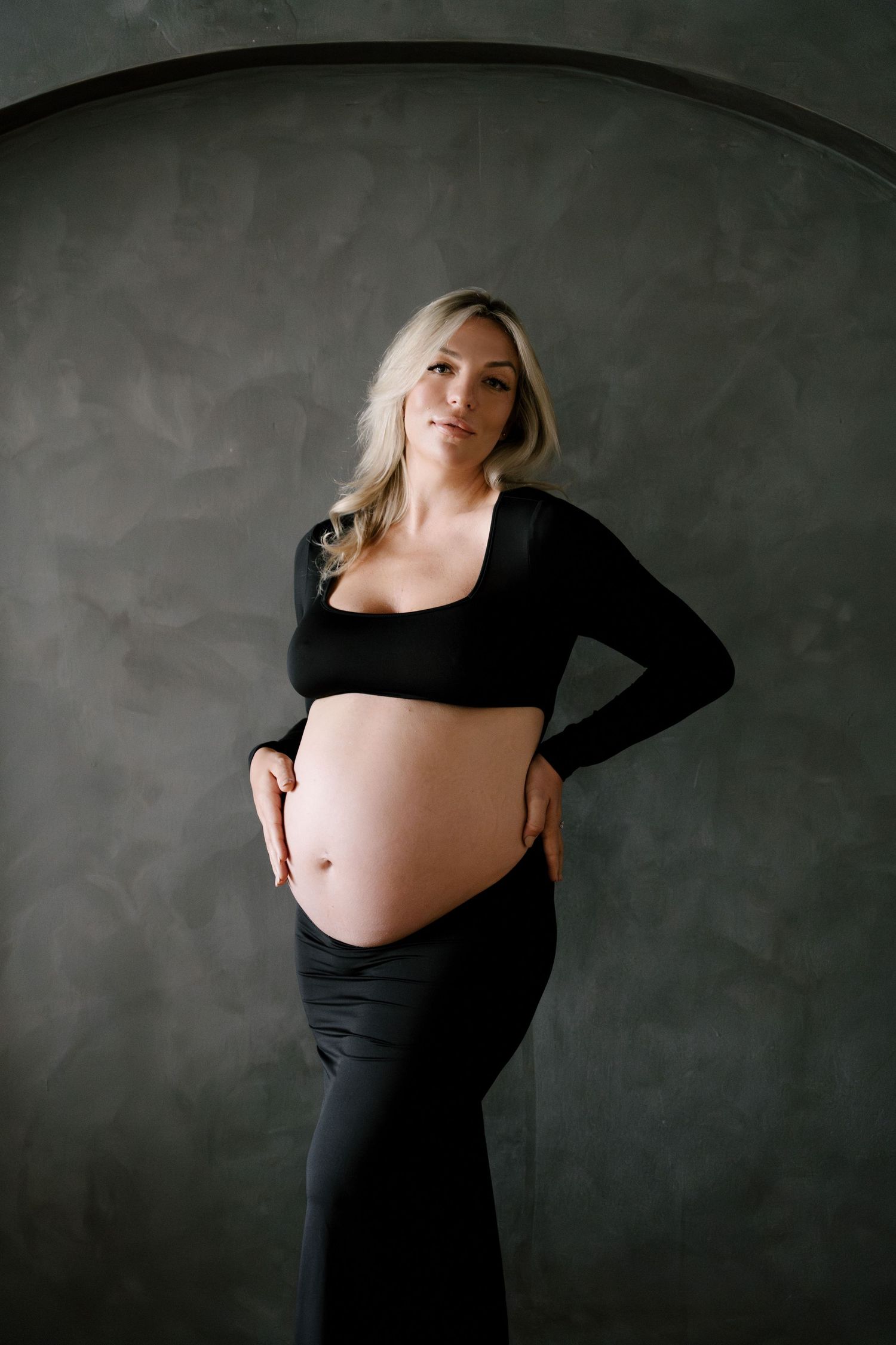 Smiling pregnant blonde woman in black crop top and skirt with hands on hips in studio setting.