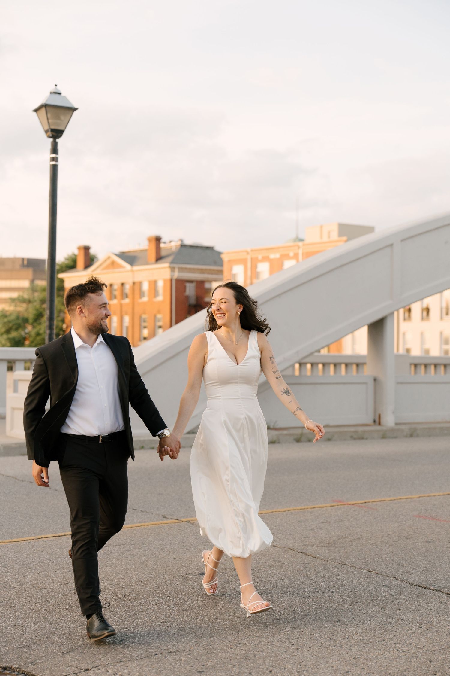 Laughing bride in white dress and groom holding hands while walking on a city street.