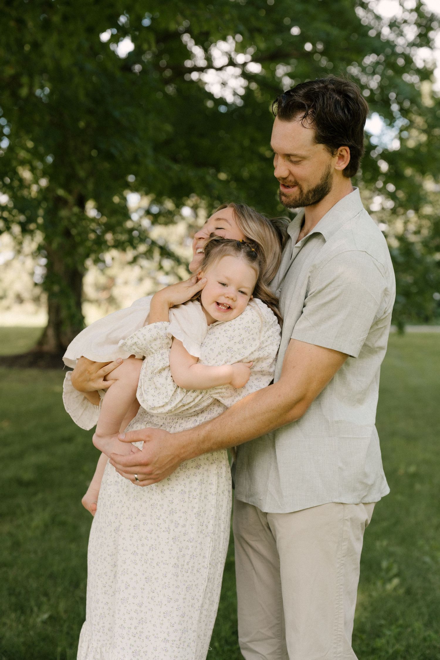 Happy family of three embracing outdoors, with a laughing toddler held between her smiling parents.