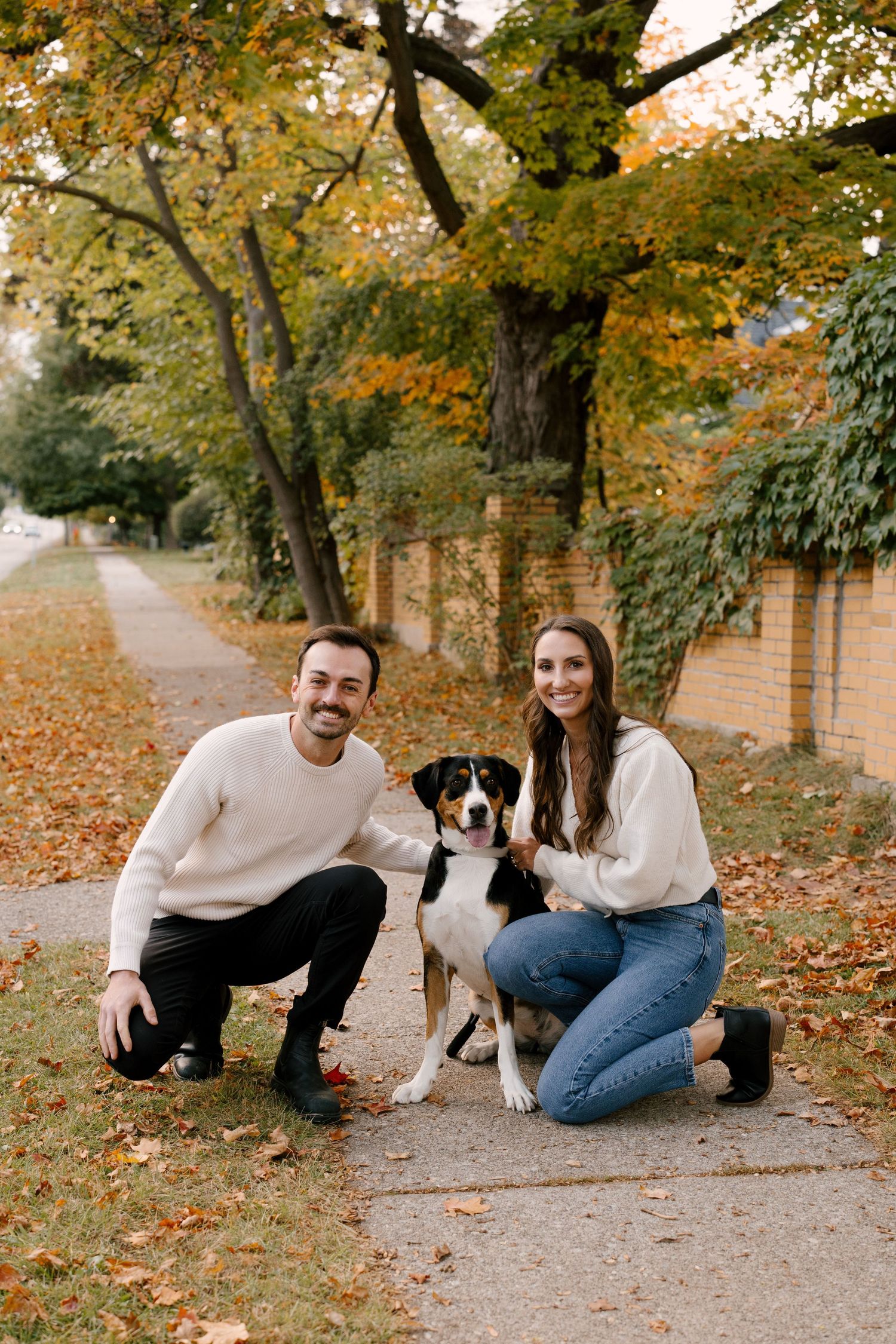 Happy couple crouching with their Bernese mix dog on a leaf-covered sidewalk in autumn.