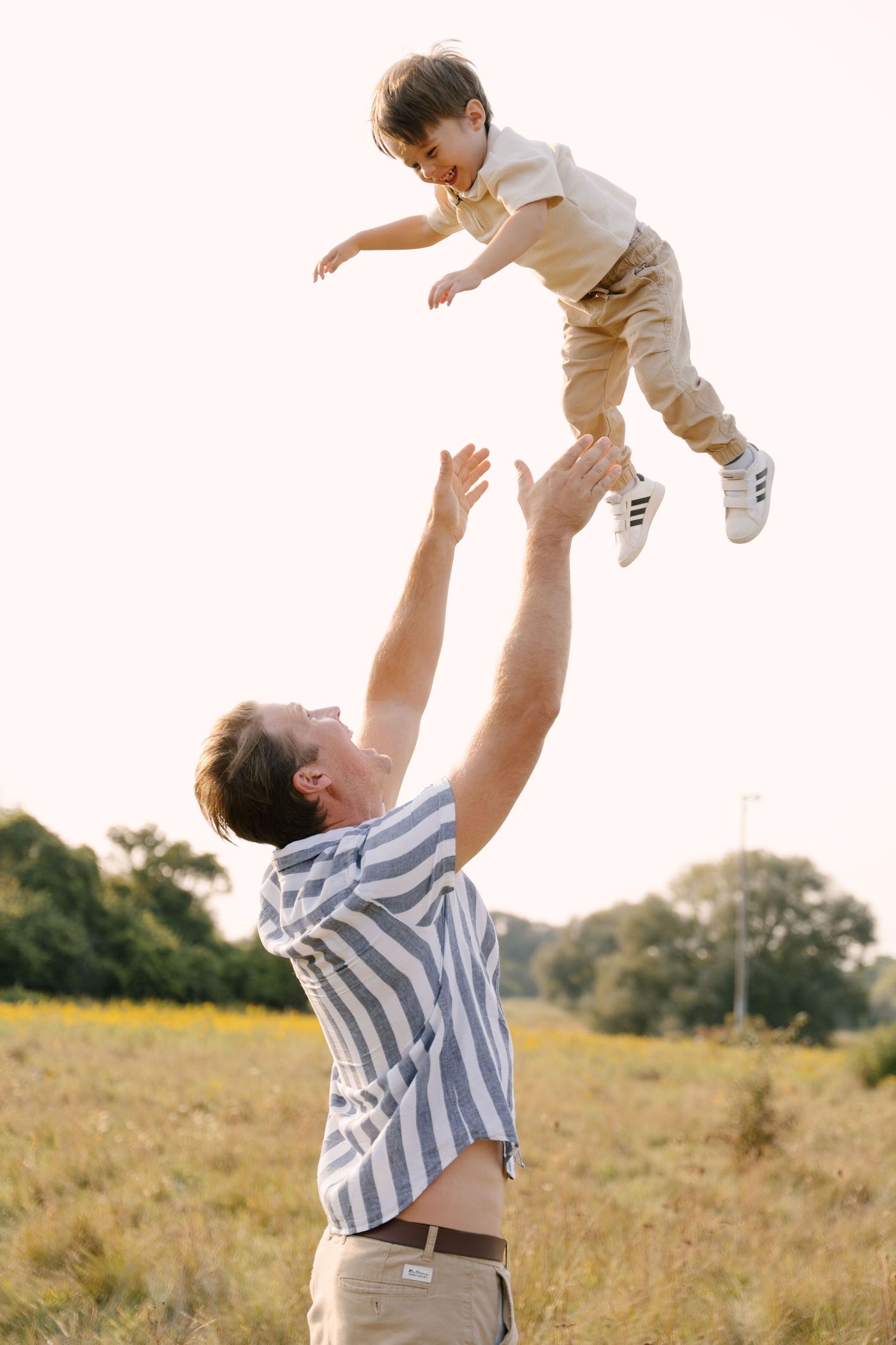 Father tossing laughing young son high into the air in a sunny field.