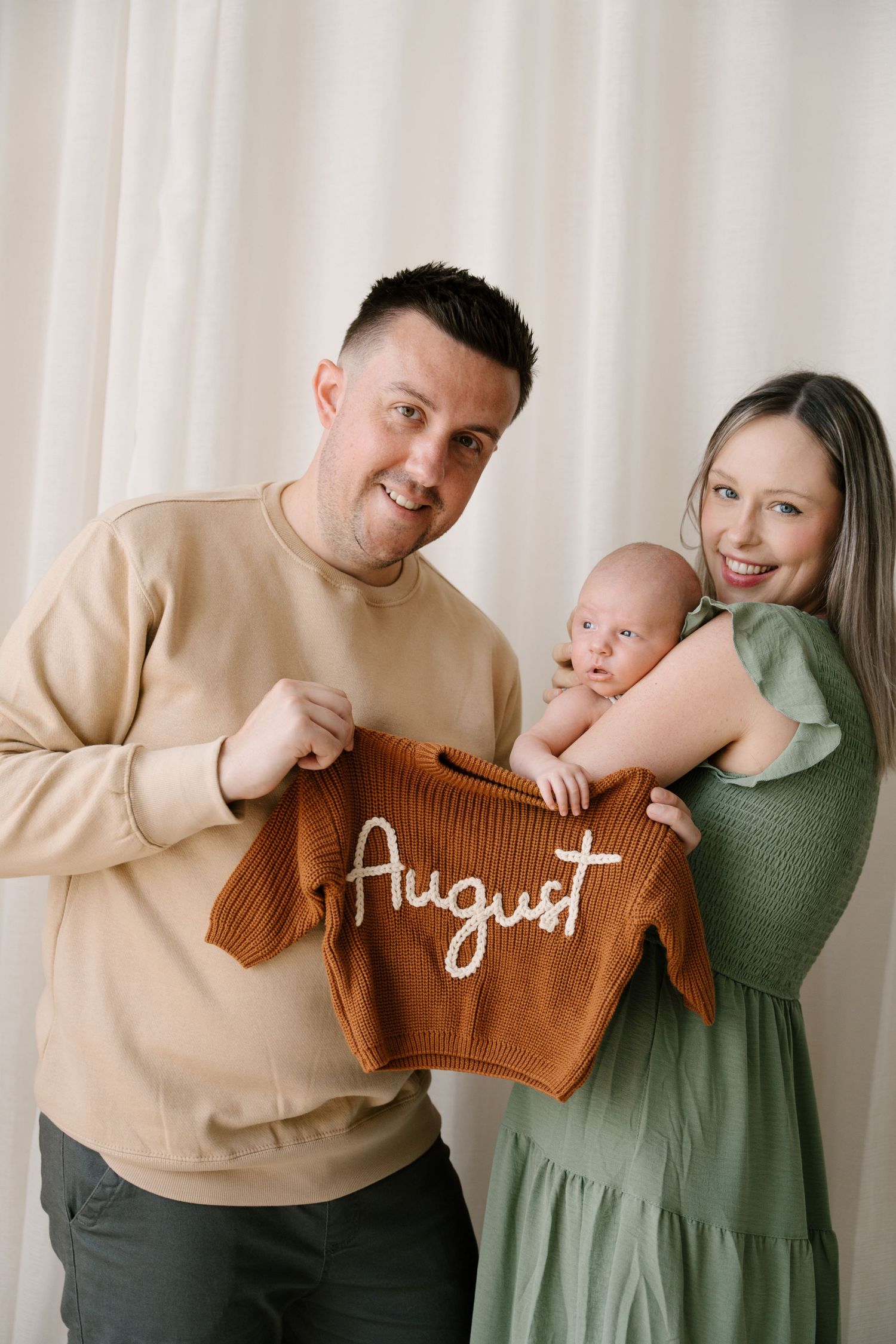Smiling parents hold their newborn baby and a brown knit onesie embroidered with the name August.