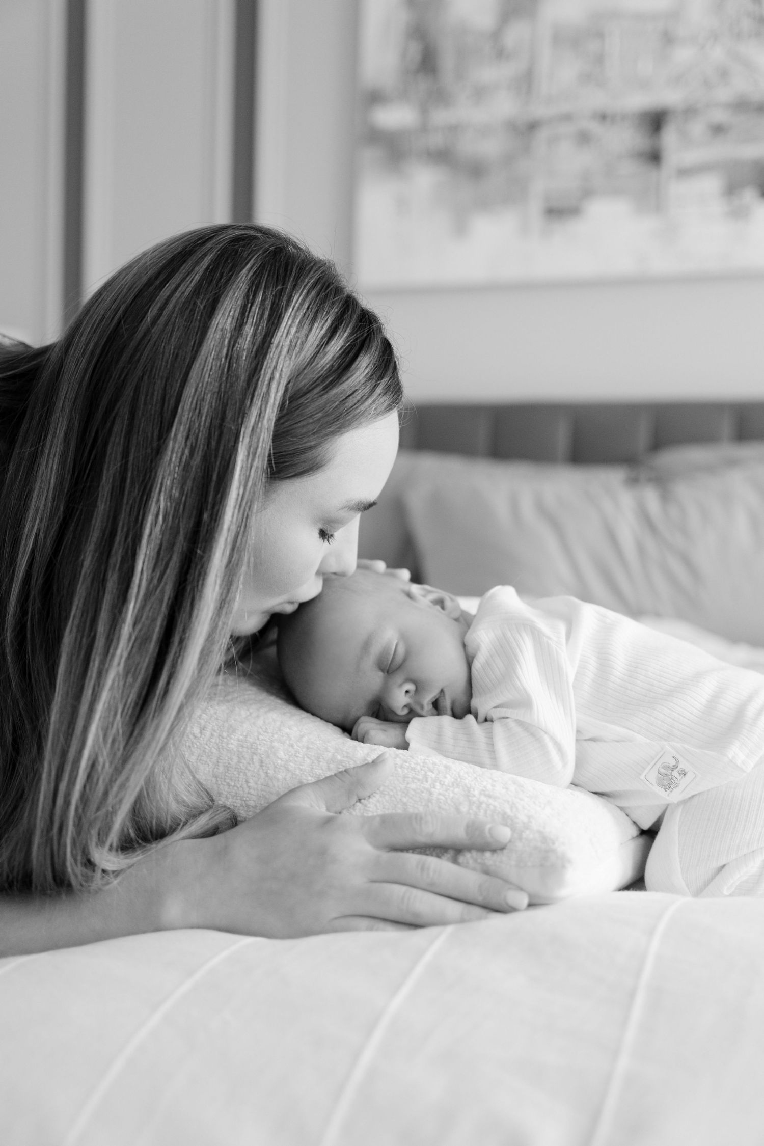 Black and white photo of a mother tenderly kissing her sleeping newborn baby on a white bed.