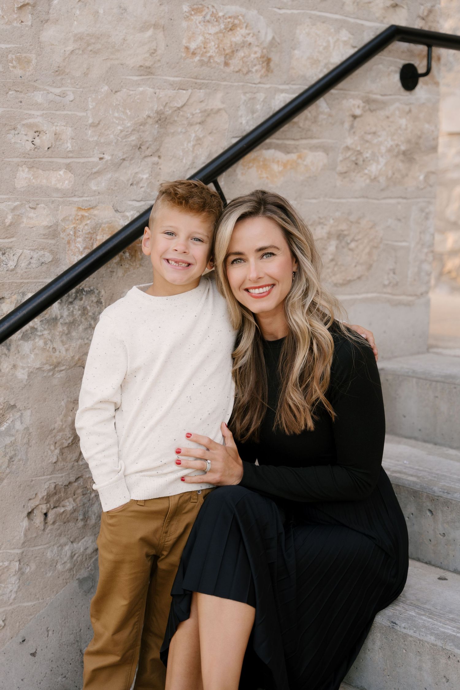 Smiling blonde mother in black dress hugging young red-haired boy on stone steps.