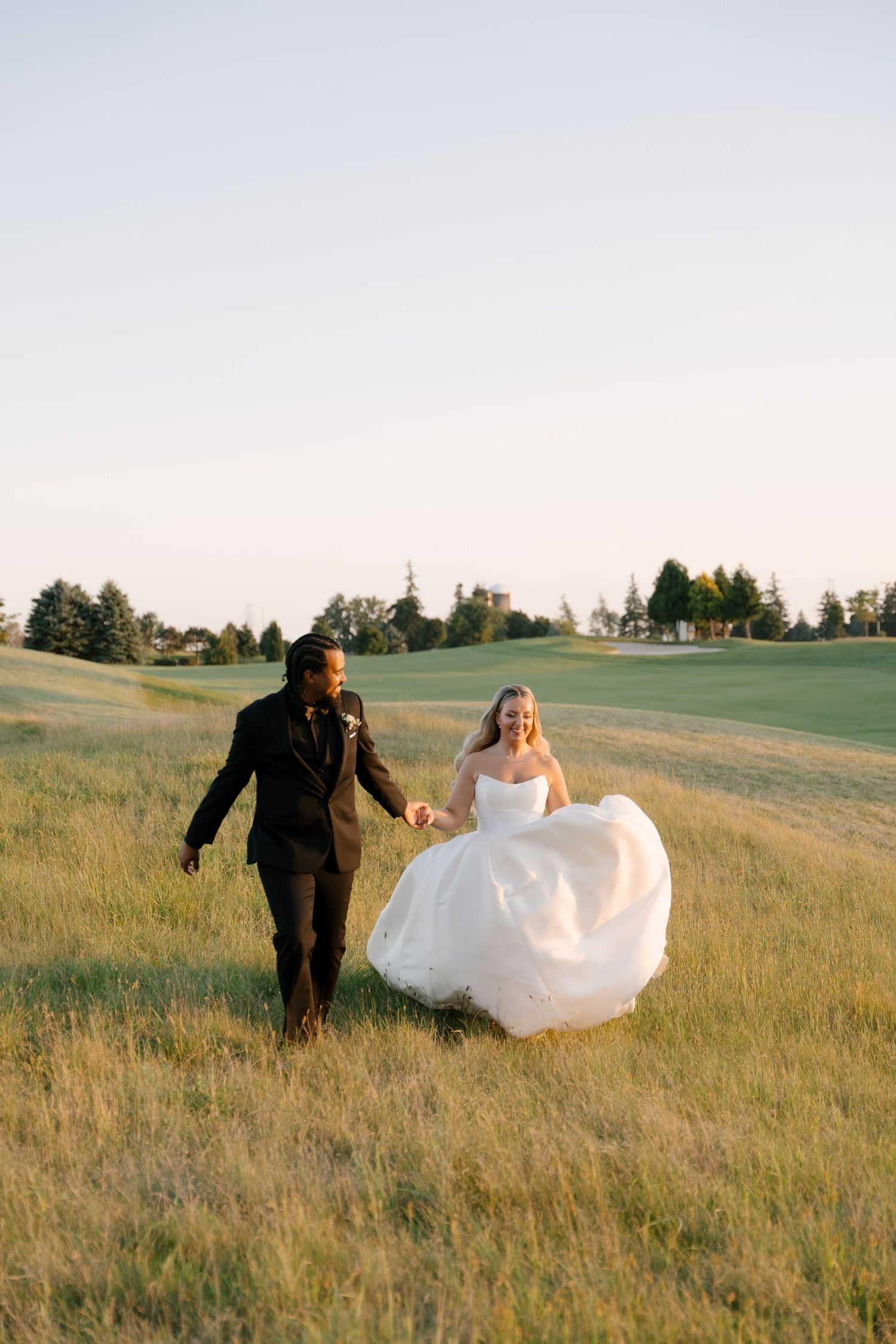 Happy bride and groom holding hands walking through golden meadow, bride in white ballgown at Whistle Bear wedding venue.