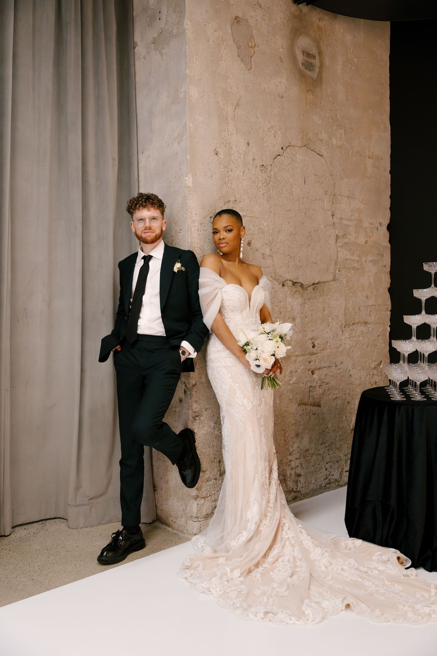 Bride in lace gown holding white bouquet stands with groom in black suit against rustic wall inside Tapestry Hall wedding venue.