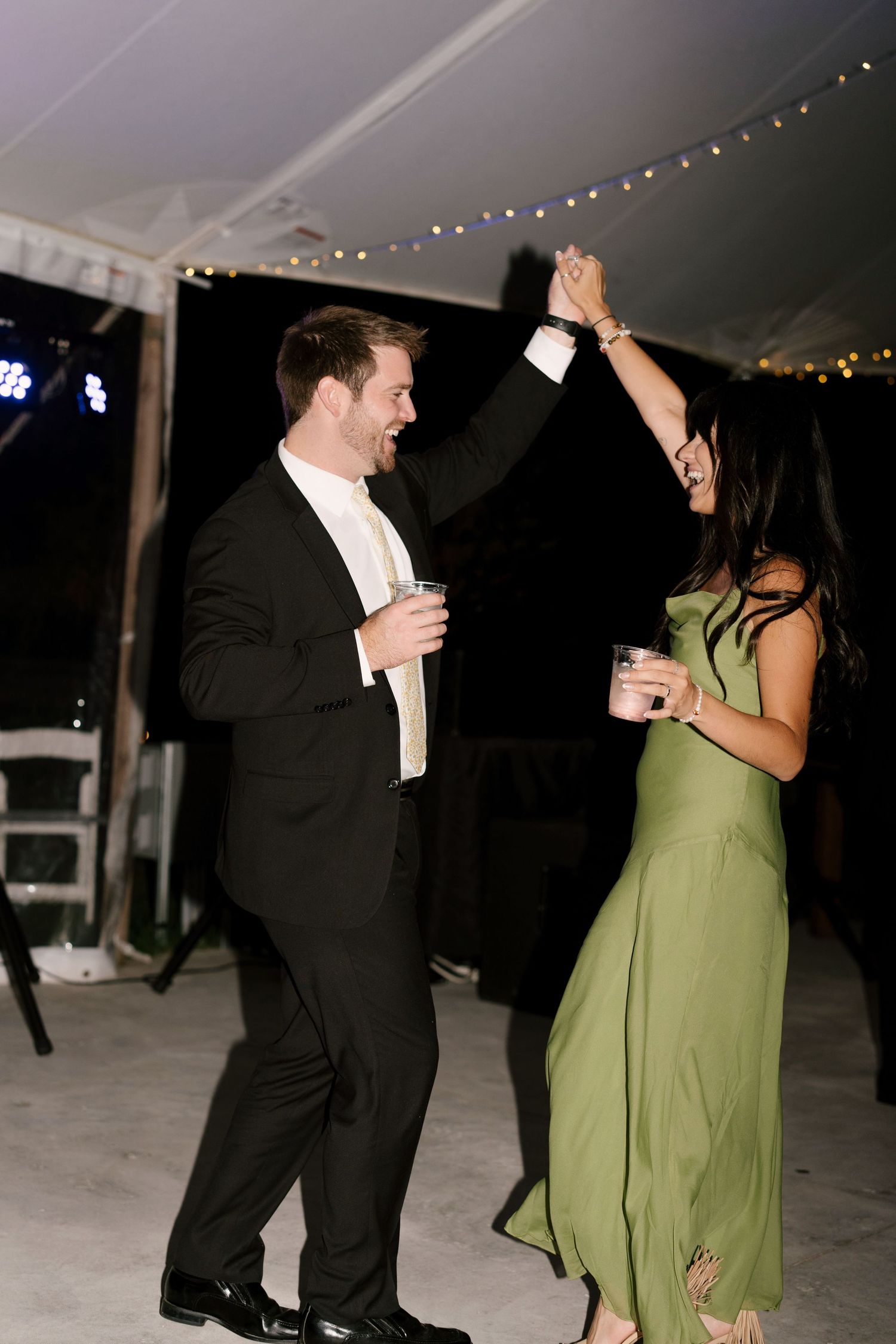 A couple dances joyfully at an outdoor evening wedding reception, the man in a black tuxedo and woman in an elegant green dress.