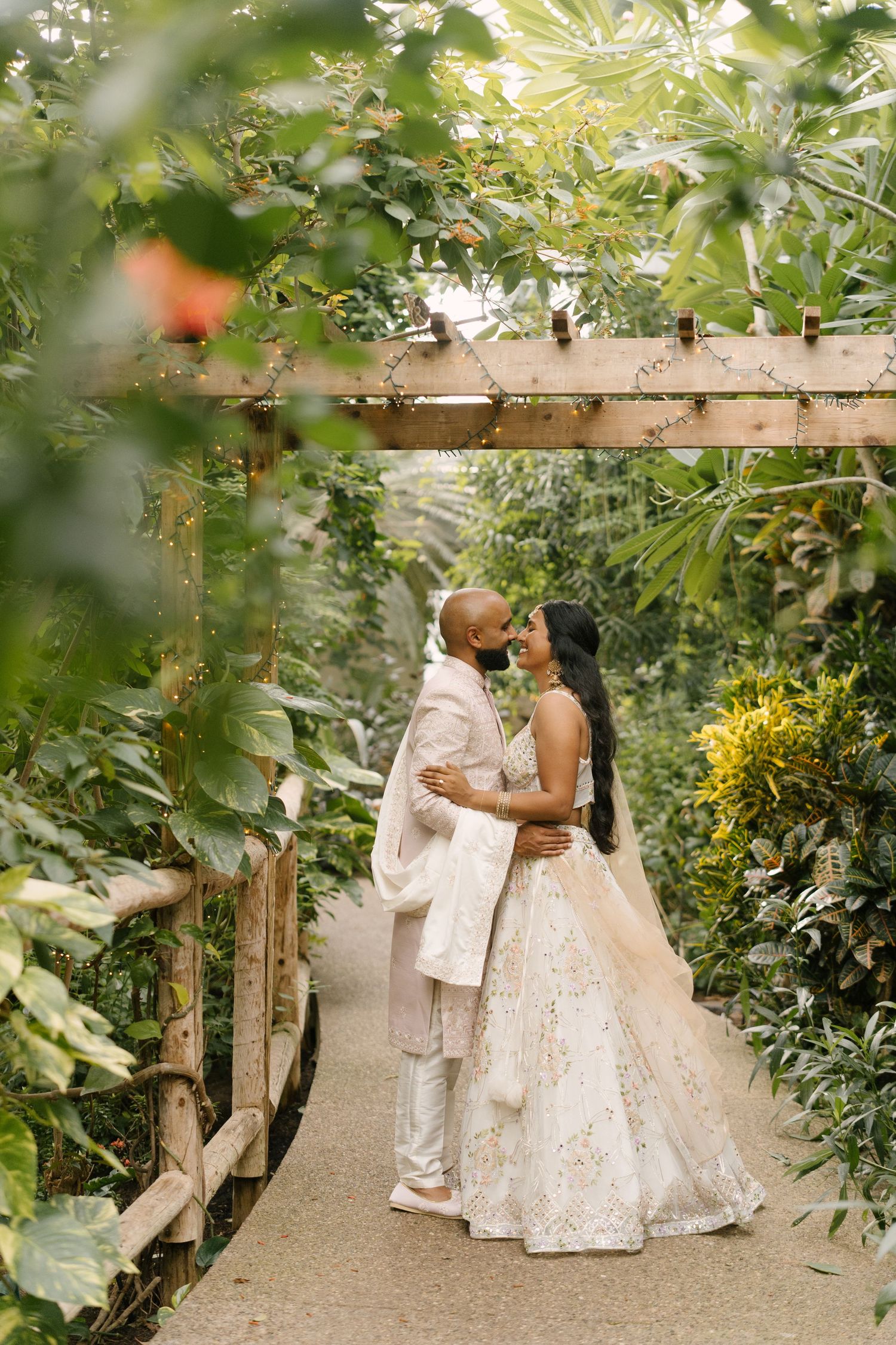 Indian couple in ivory wedding attire embrace on garden path beneath lush green pergola.