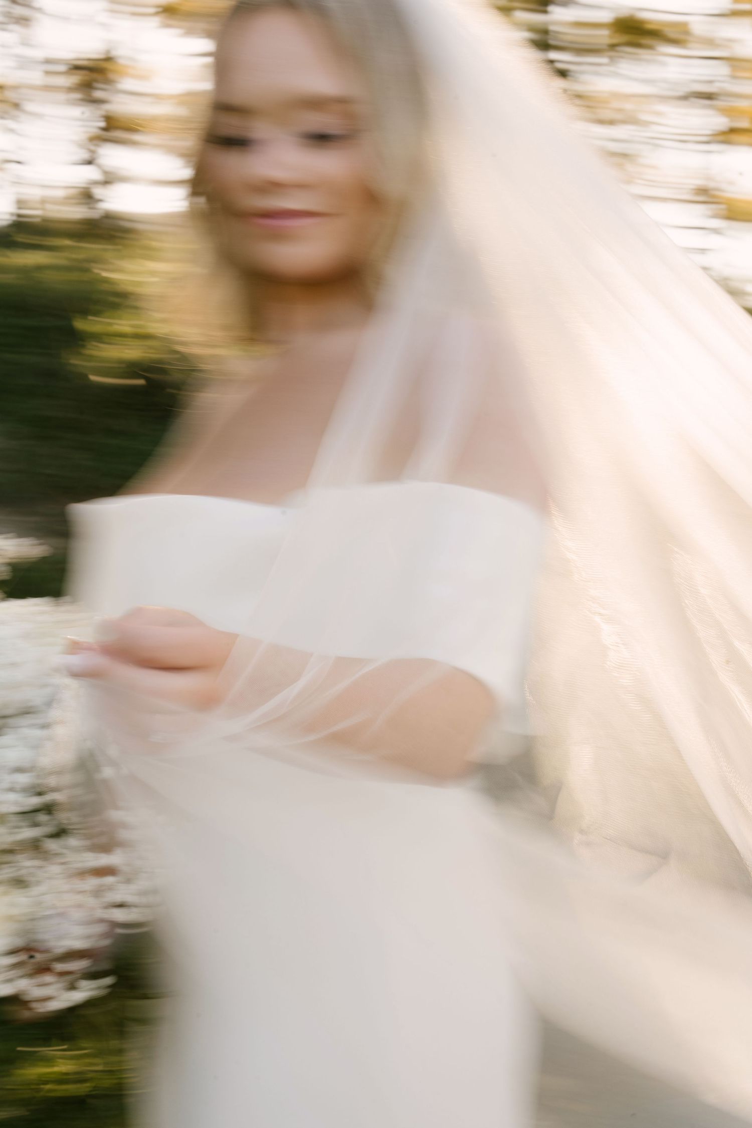 Artistic motion-blur photo of blonde bride in white off-shoulder gown and veil outdoors.