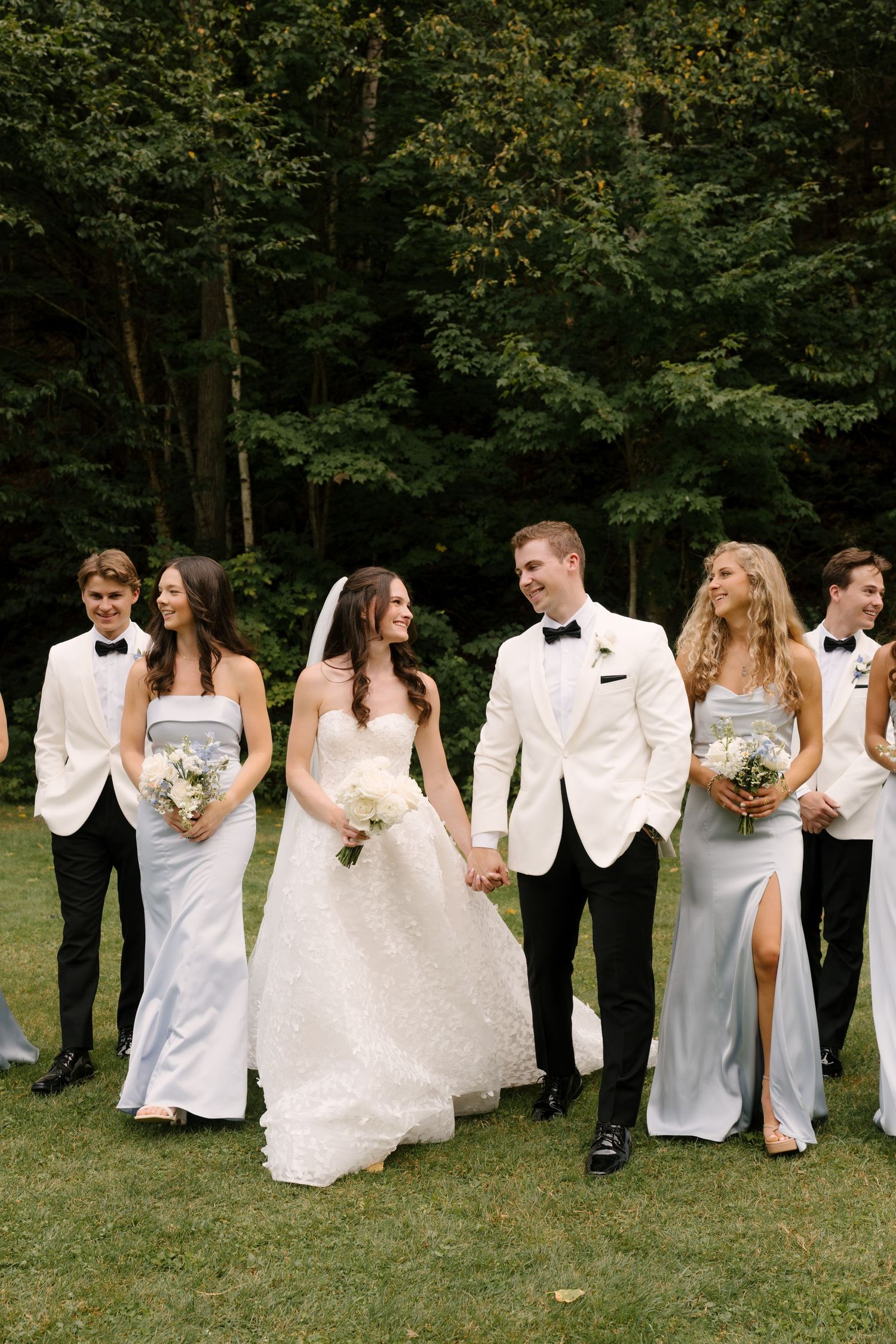 Wedding party walks outdoors in white tuxedos and light blue bridesmaid dresses, bride in white gown holds bouquet.