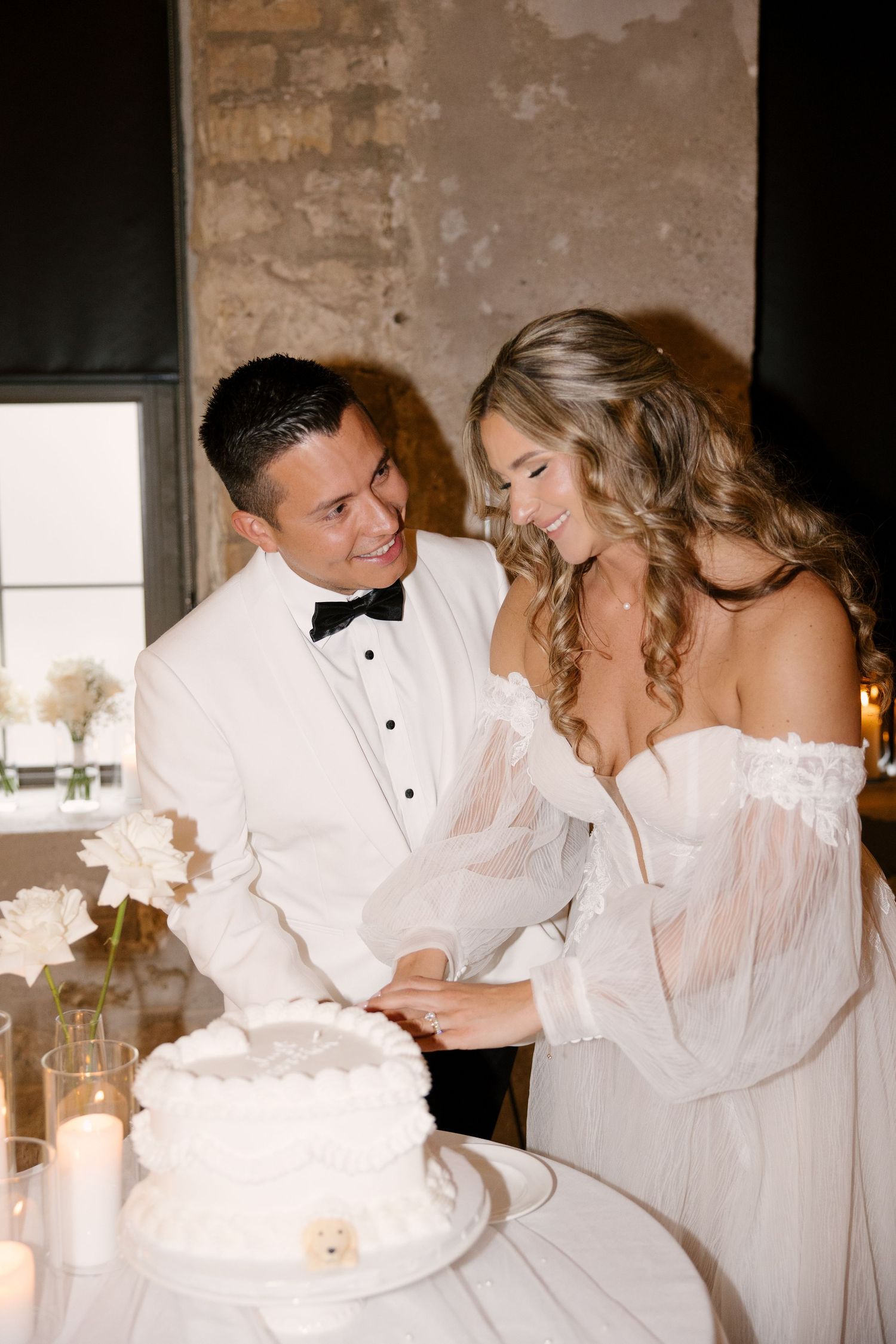 Smiling bride and groom cutting white wedding cake together by candlelight at reception in Tapestry Hall wedding venue.