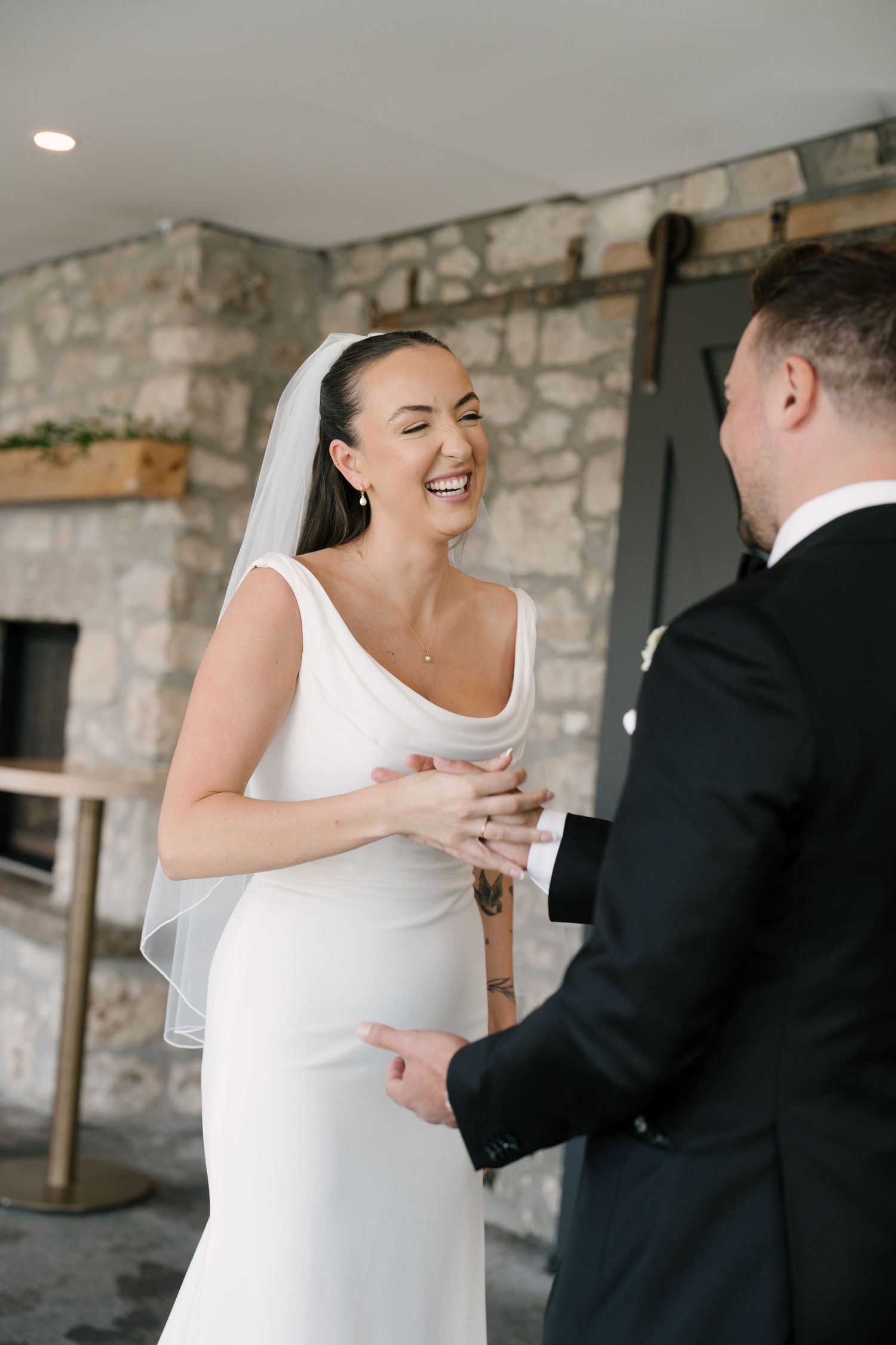 Bride in white gown and veil laughs joyfully while holding groom's hands during wedding first look at Cambridge Mill.
