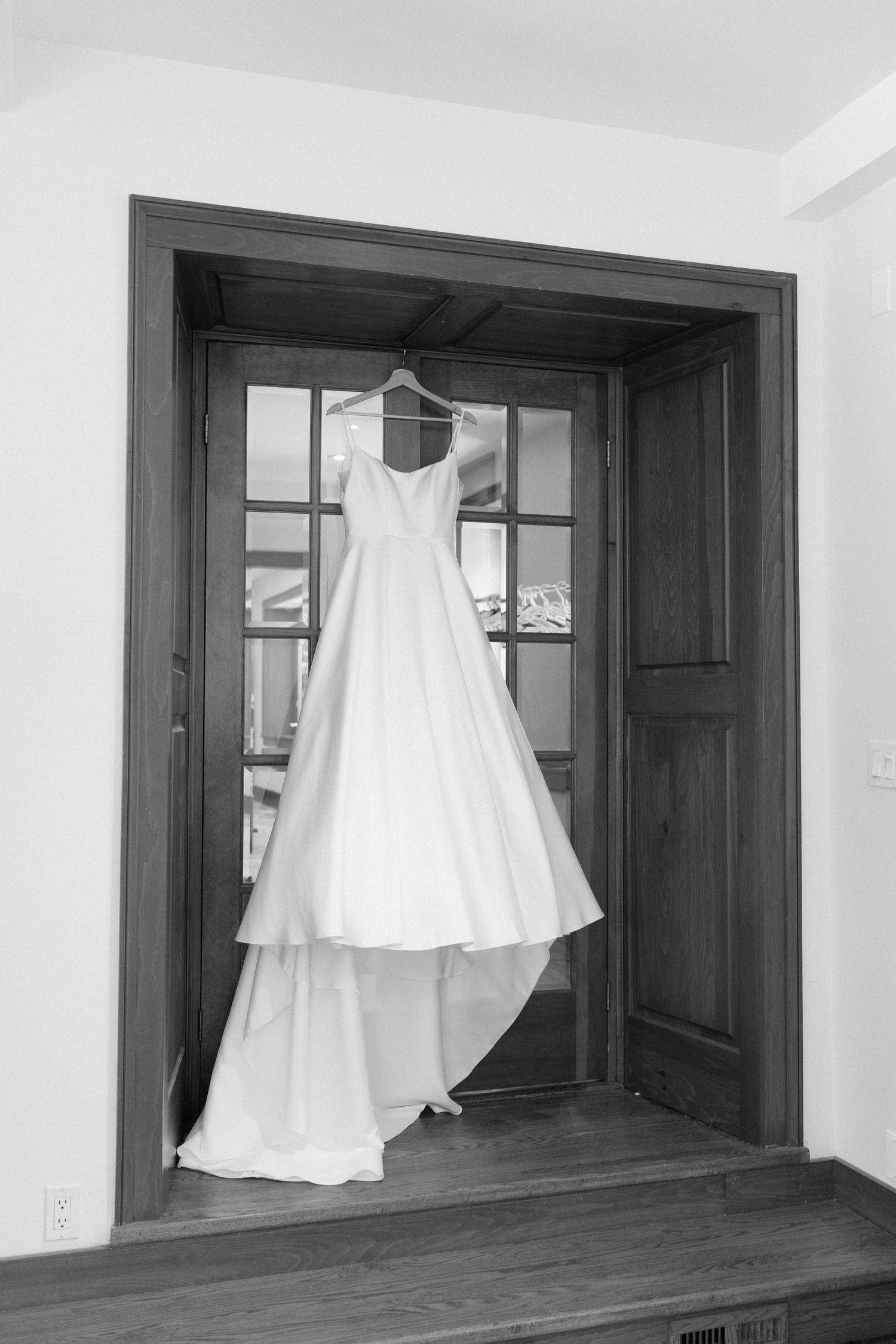 Elegant white wedding gown hanging on wooden doorframe in classic black and white bridal preparation photo.