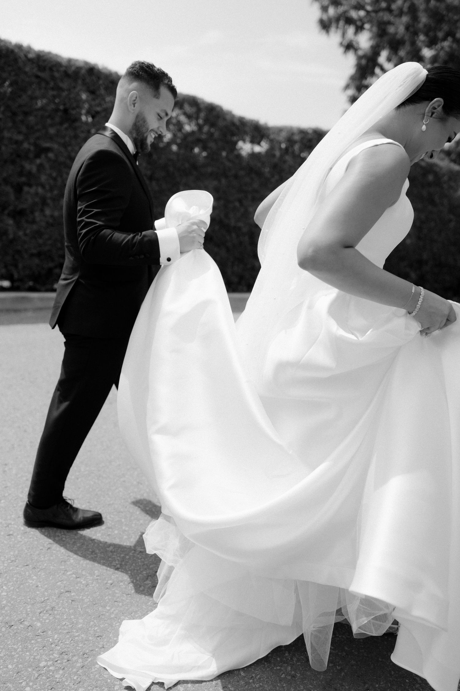 Black and white photo of groom holding bride's wedding dress train outdoors near hedge.