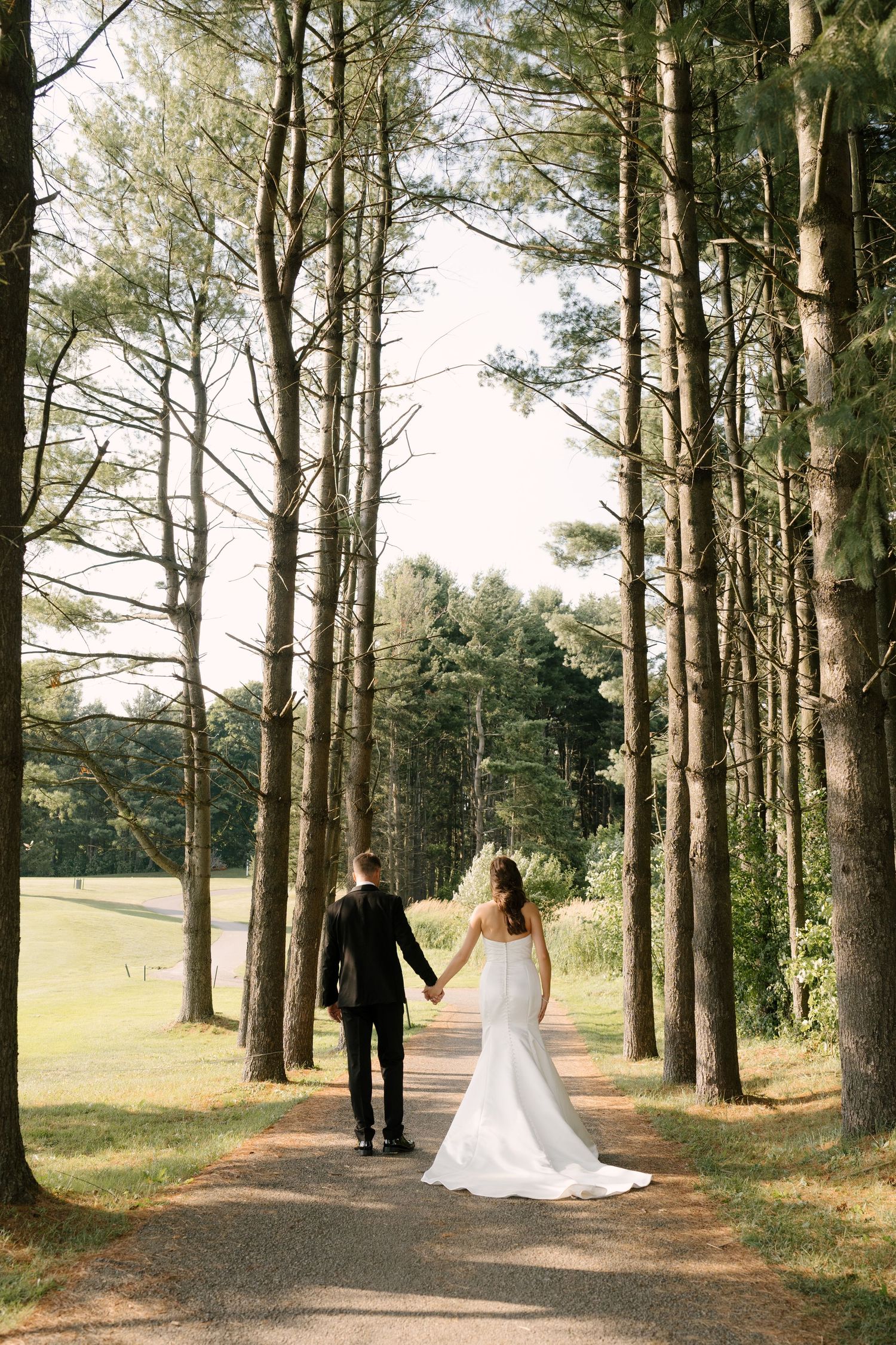 Bride and groom walk hand in hand down a serene tree-lined path, bride in a fitted white mermaid wedding gown.