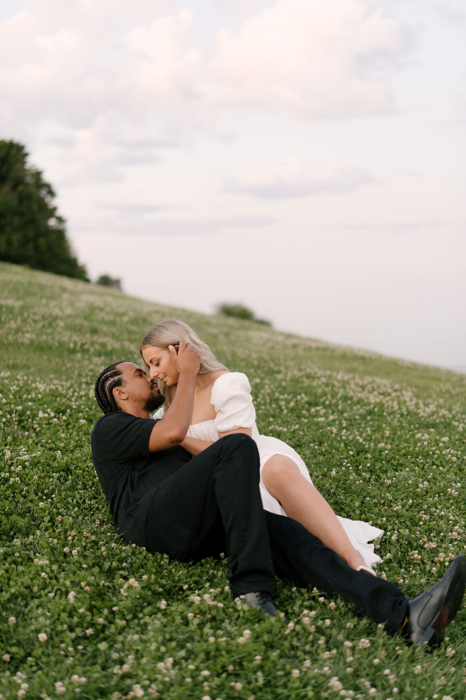 Couple sharing a romantic kiss while sitting in a lush green meadow filled with wildflowers at dusk.