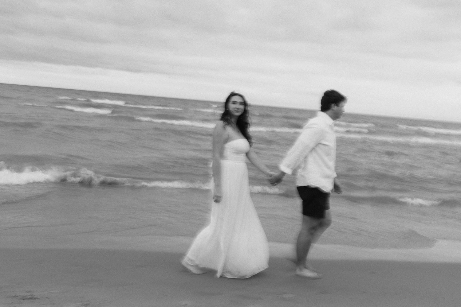 Black and white photo of bride in strapless gown and groom holding hands walking along beach waves.