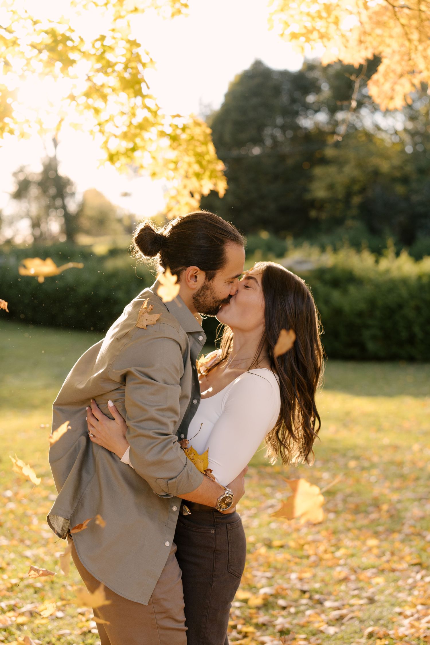 Couple sharing a romantic kiss in a sunlit autumn park, surrounded by golden falling leaves and lush green trees.