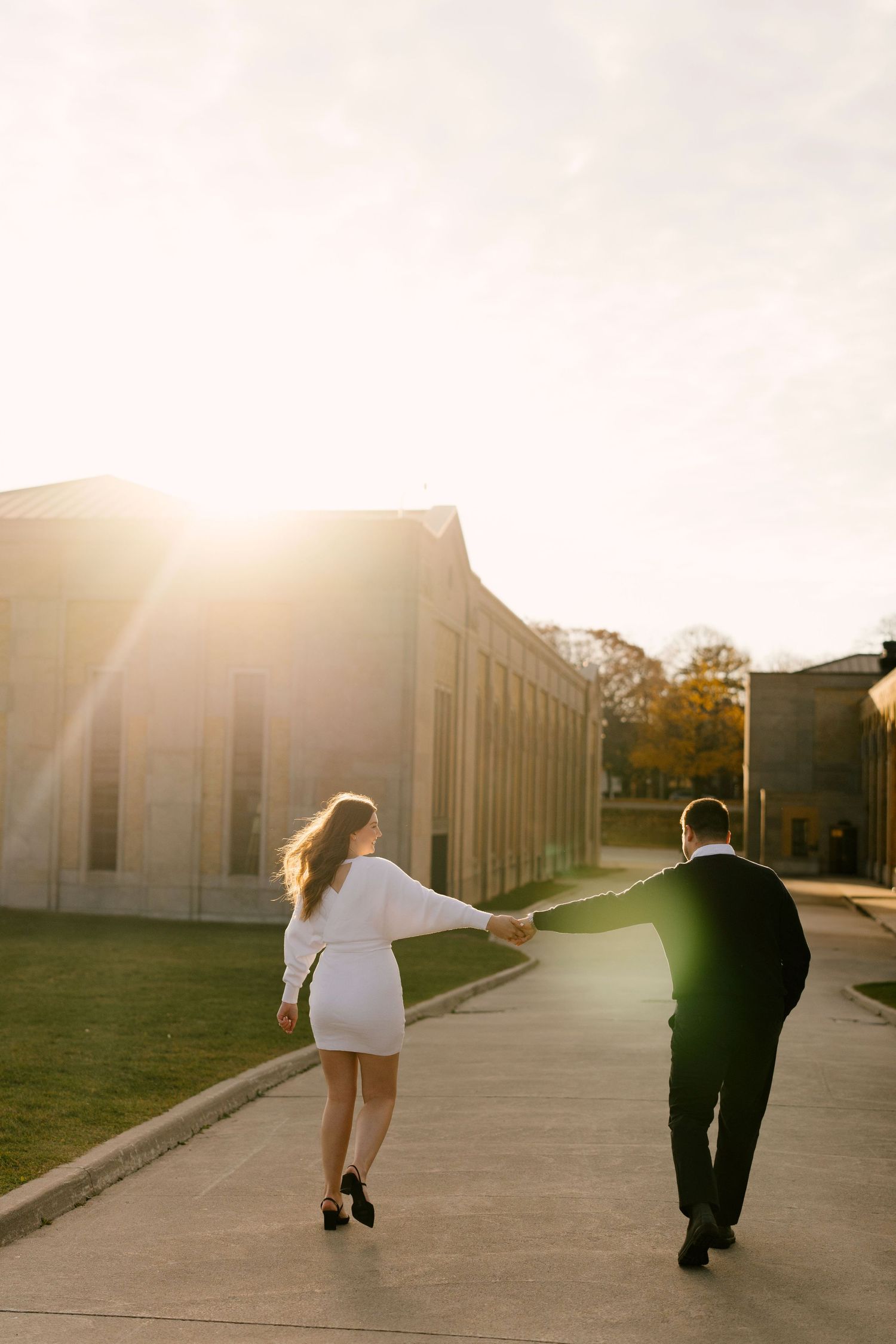 Couple walking hand in hand toward golden sunset light beside a large stone building.