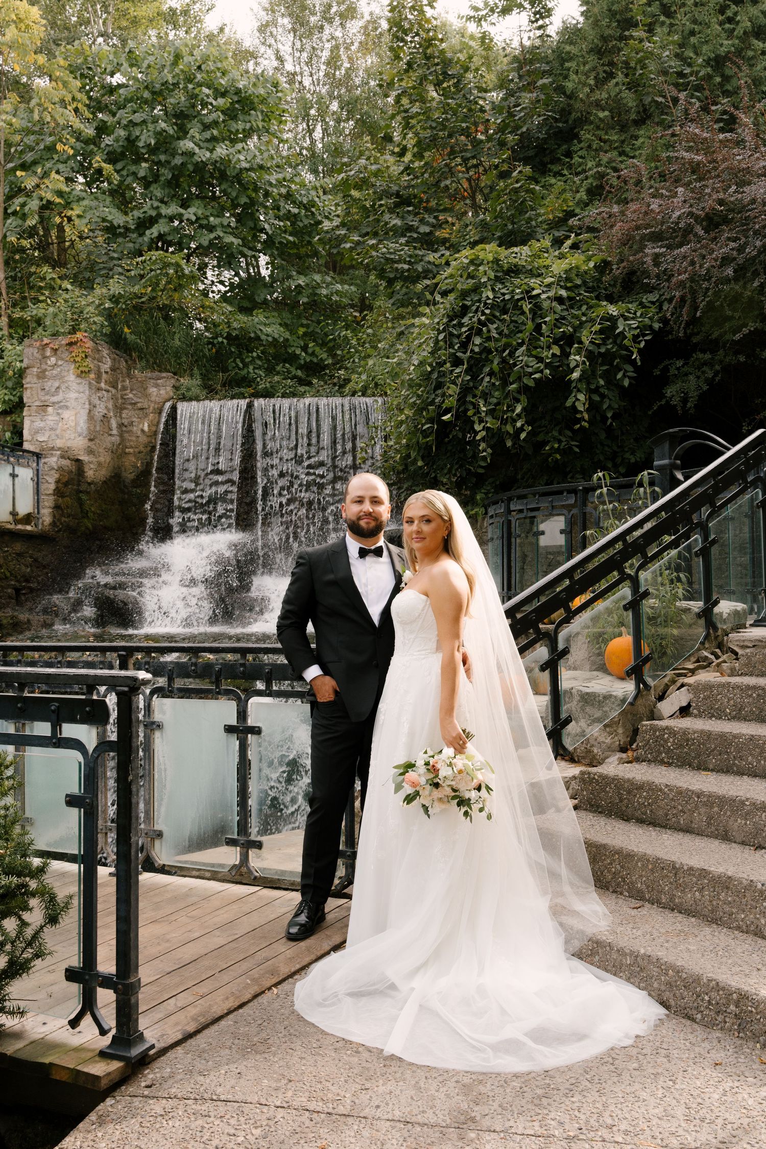 Bride and groom standing together near a waterfall backdrop, bride holding bouquet with long flowing veil at Ancaster Mill.