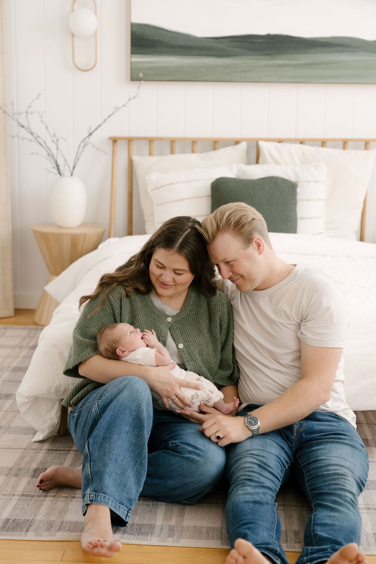 New parents sitting on bedroom floor smiling at their newborn baby wrapped in white swaddle blanket.