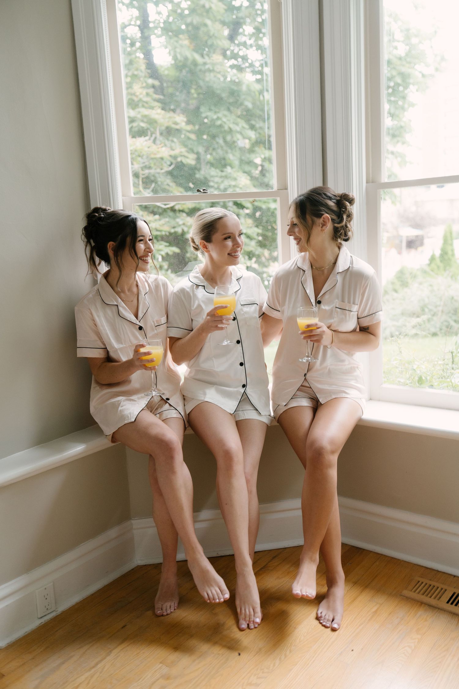 Three bridesmaids in matching white pajamas sit by a bright window, smiling and holding glasses of orange juice.