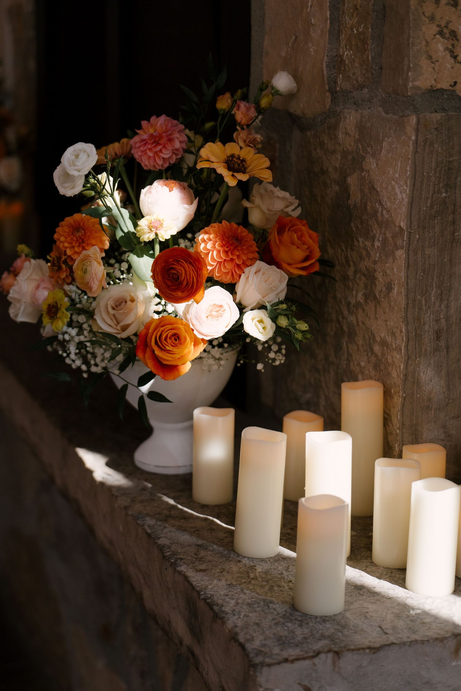 White vase with orange, pink, and cream flowers beside ivory pillar candles on a stone ledge.