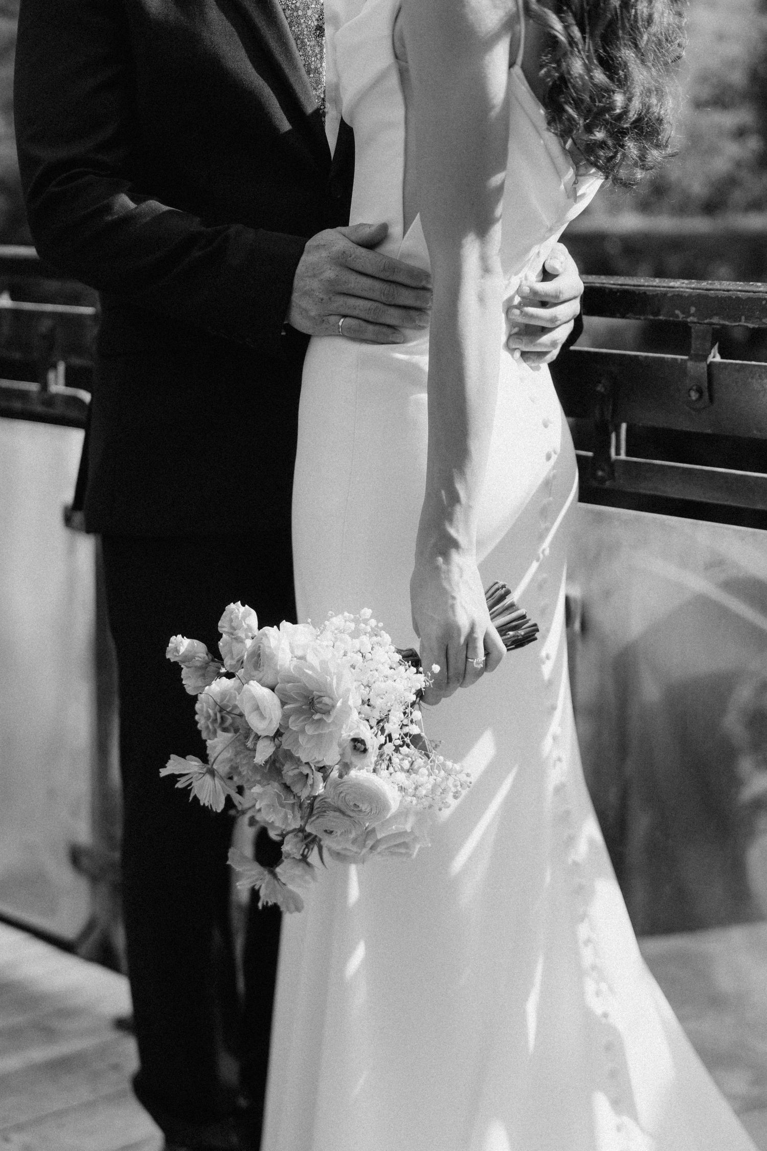 Black and white photo of groom embracing bride holding a lush bridal bouquet on a bridge.