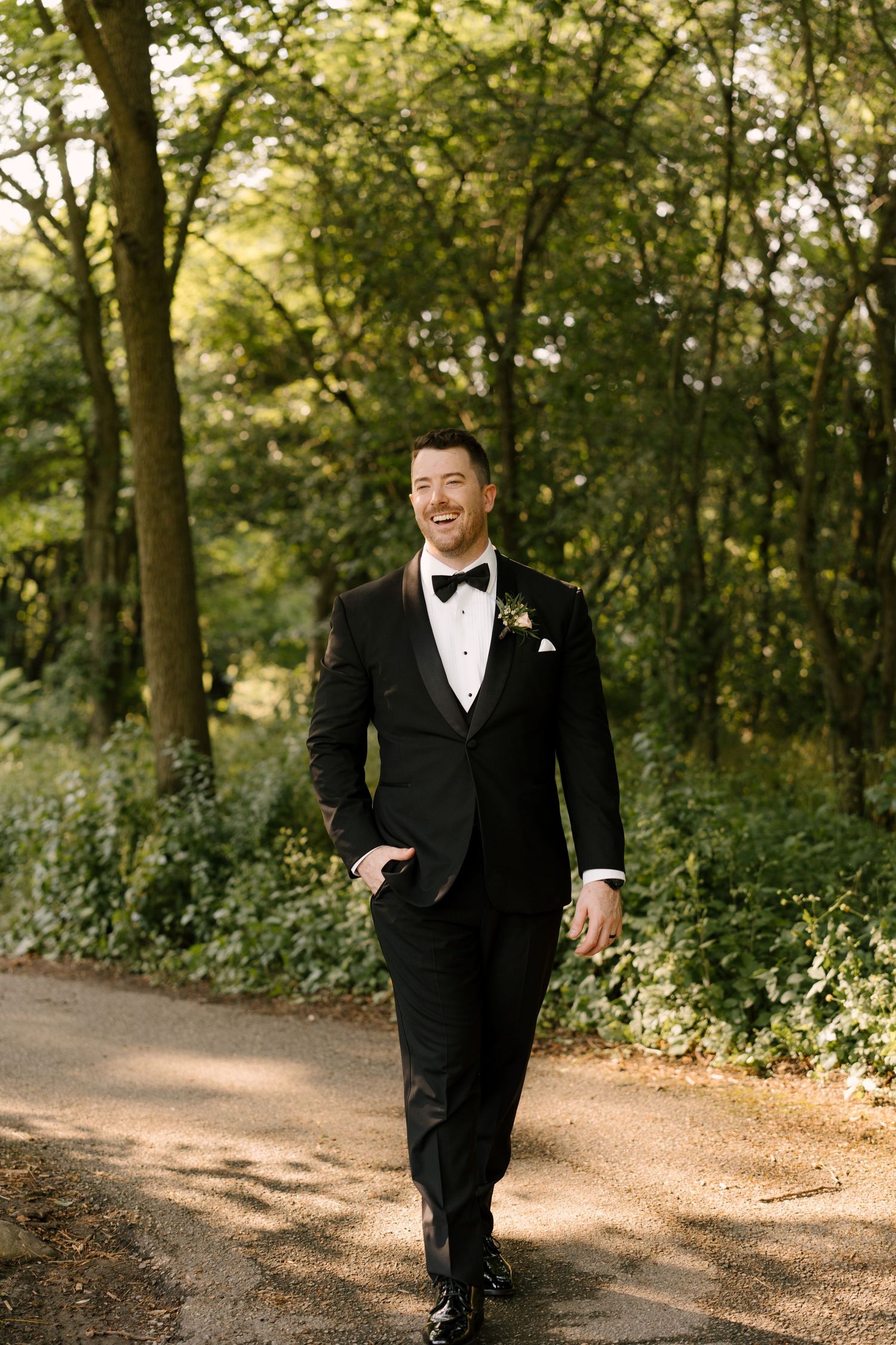 Smiling groom in black tuxedo with bow tie walks along a tree-lined path on his wedding day at St Marys Golf Club.