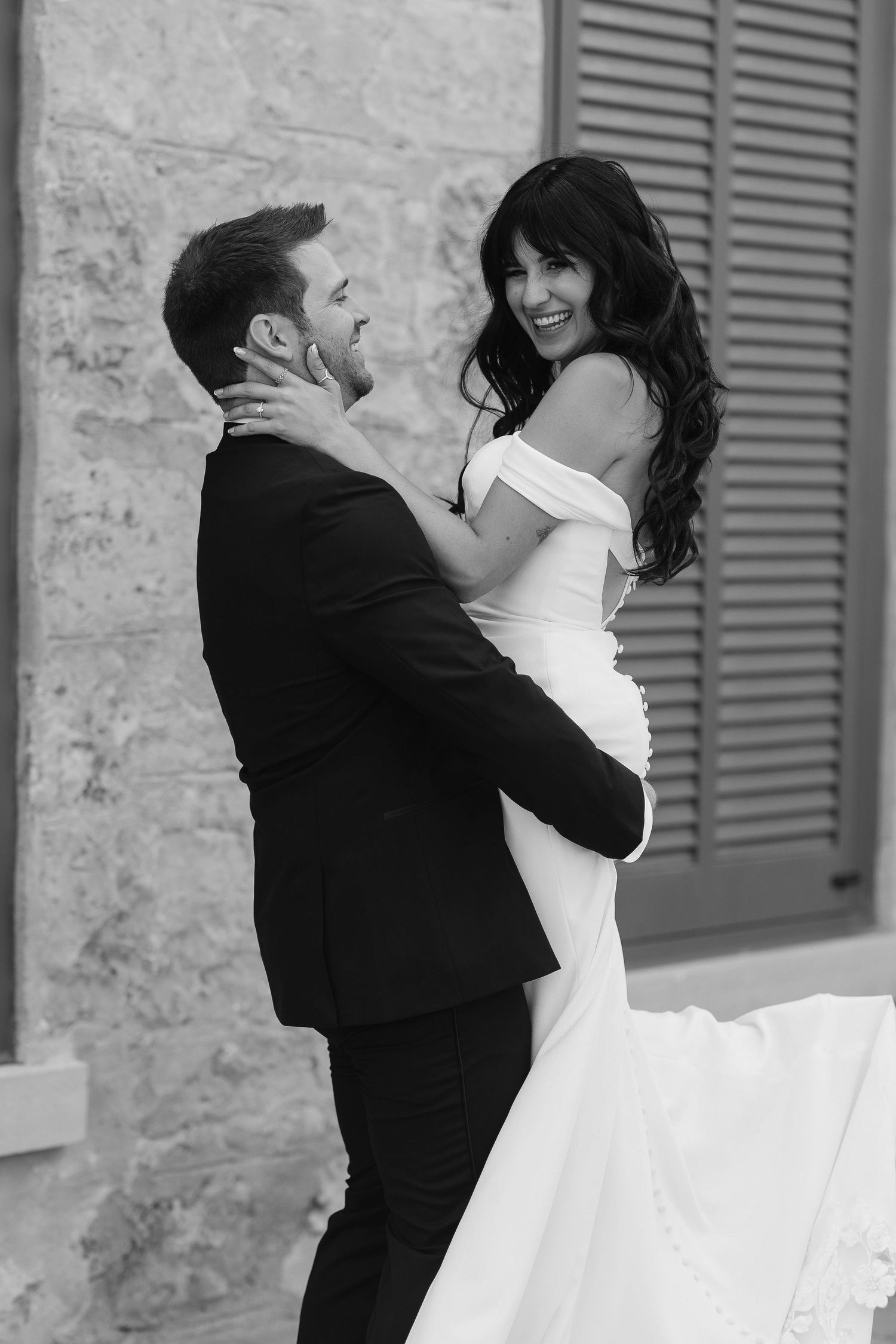 Black and white photo of a groom lifting his smiling bride in a fitted off-shoulder wedding gown outside Tapestry Hall.