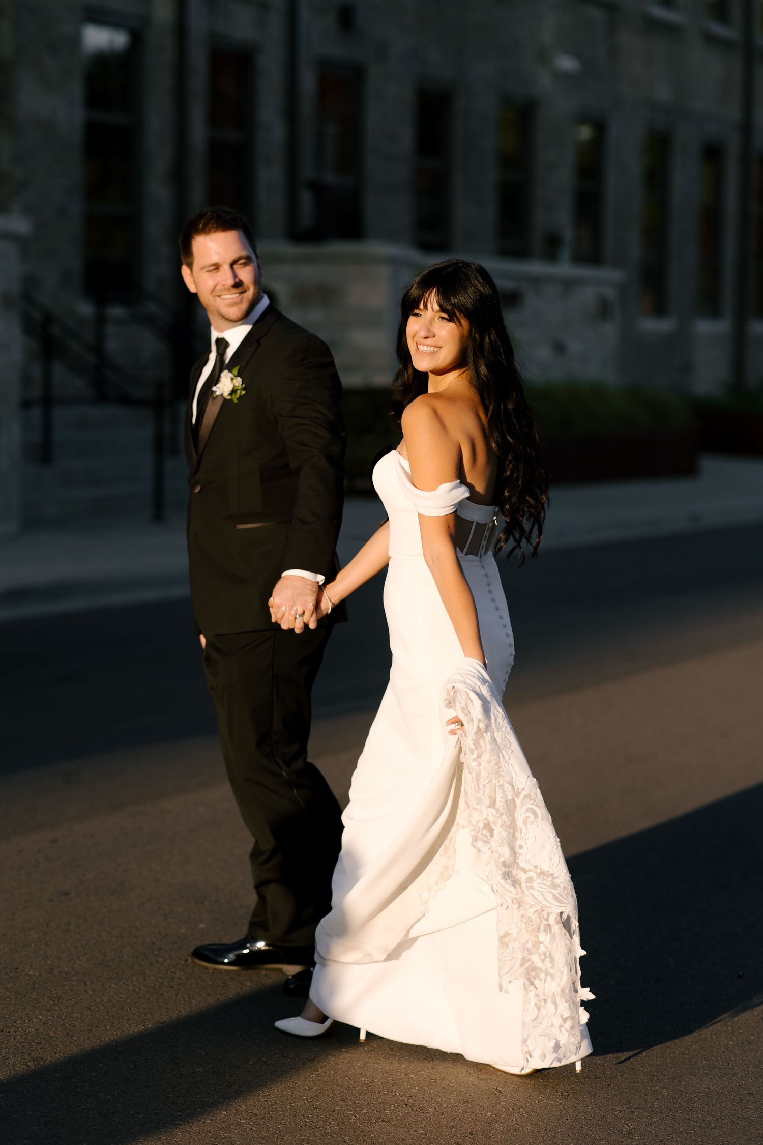 Bride and groom holding hands walking outdoors in golden sunset light, she in white gown, he in black tux.