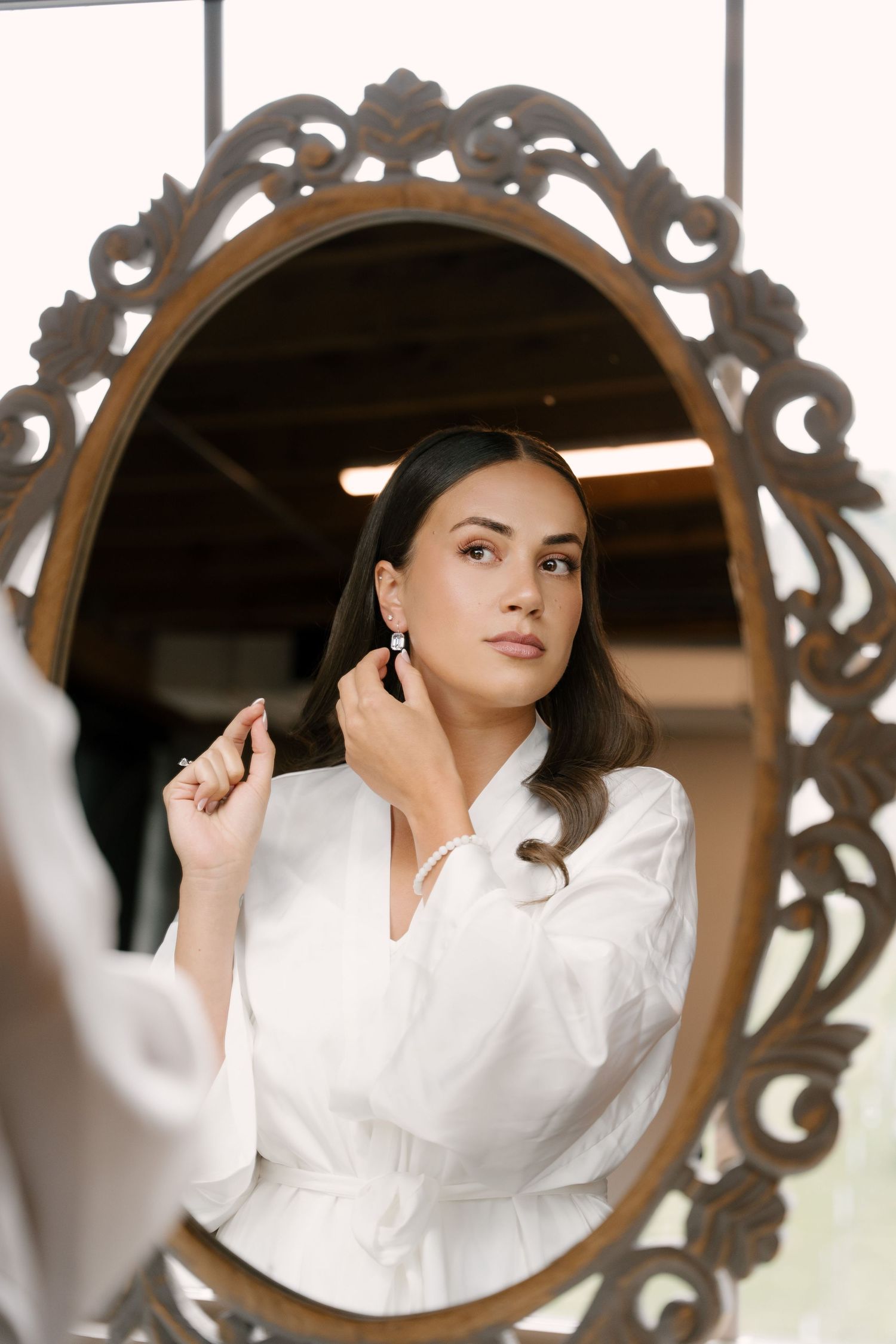 Bride in white robe putting on earrings while gazing into ornate vintage oval mirror during wedding preparations.