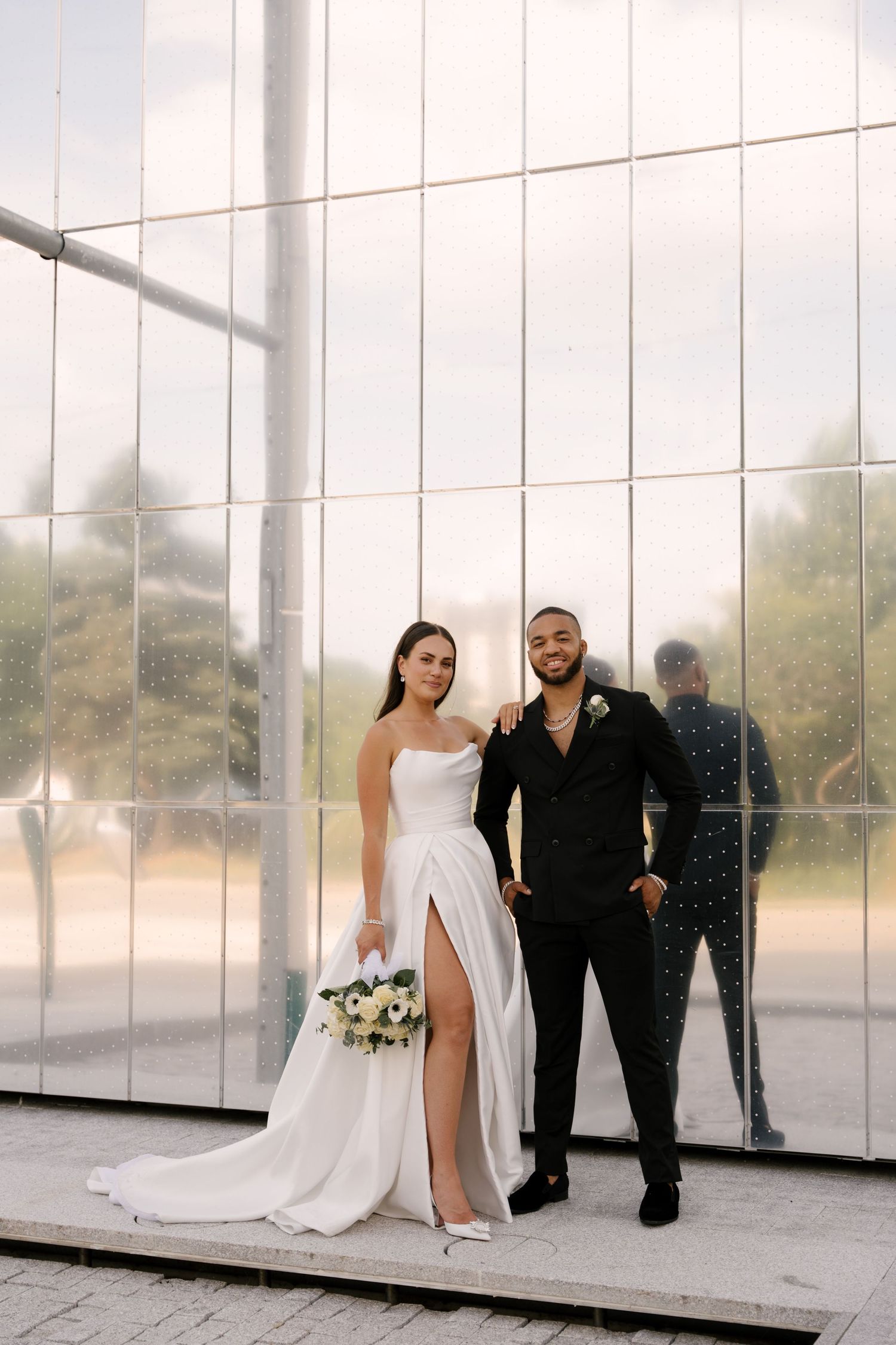 Bride in white slit gown holding bouquet poses with groom in black suit against modern glass wall outside Tapestry Hall wedding venue.