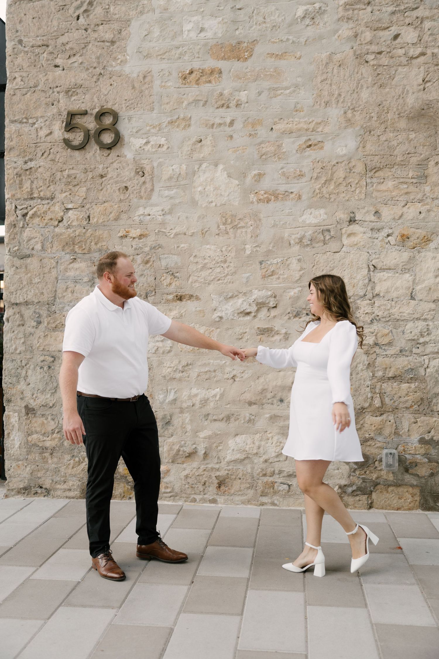 Couple holding hands and dancing playfully in front of a stone wall marked with number 58.