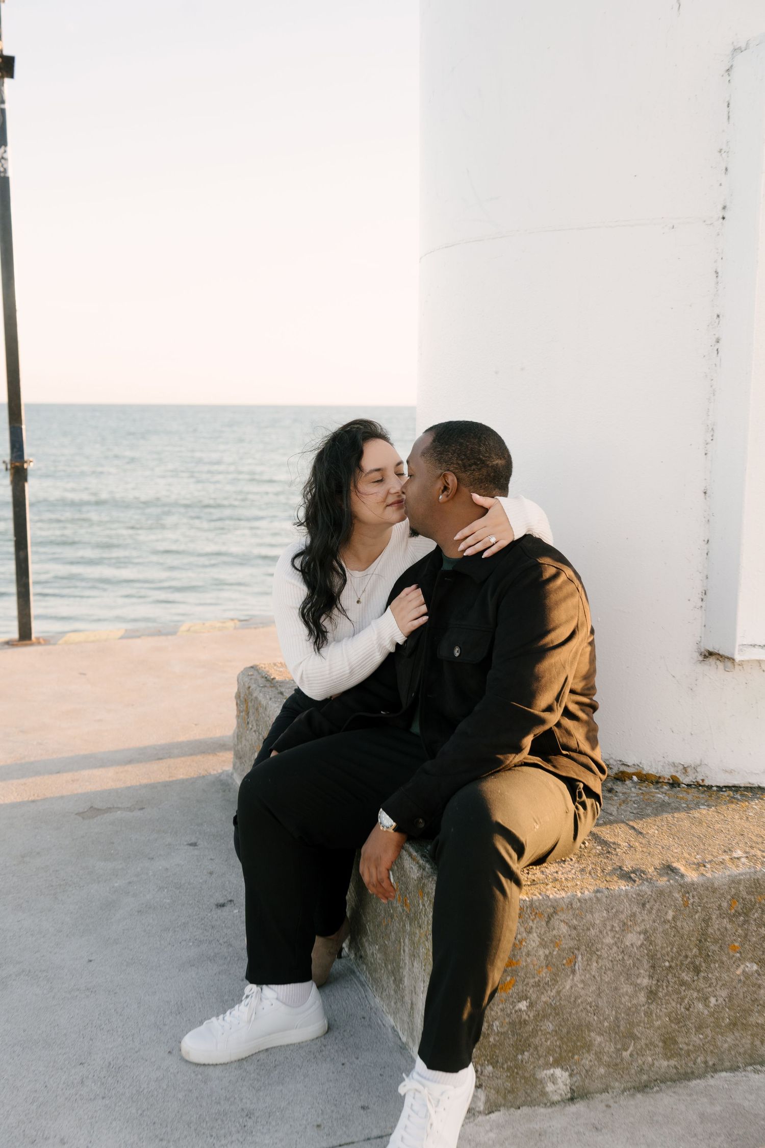 Couple sharing a kiss while seated on a waterfront ledge at sunset by the ocean.