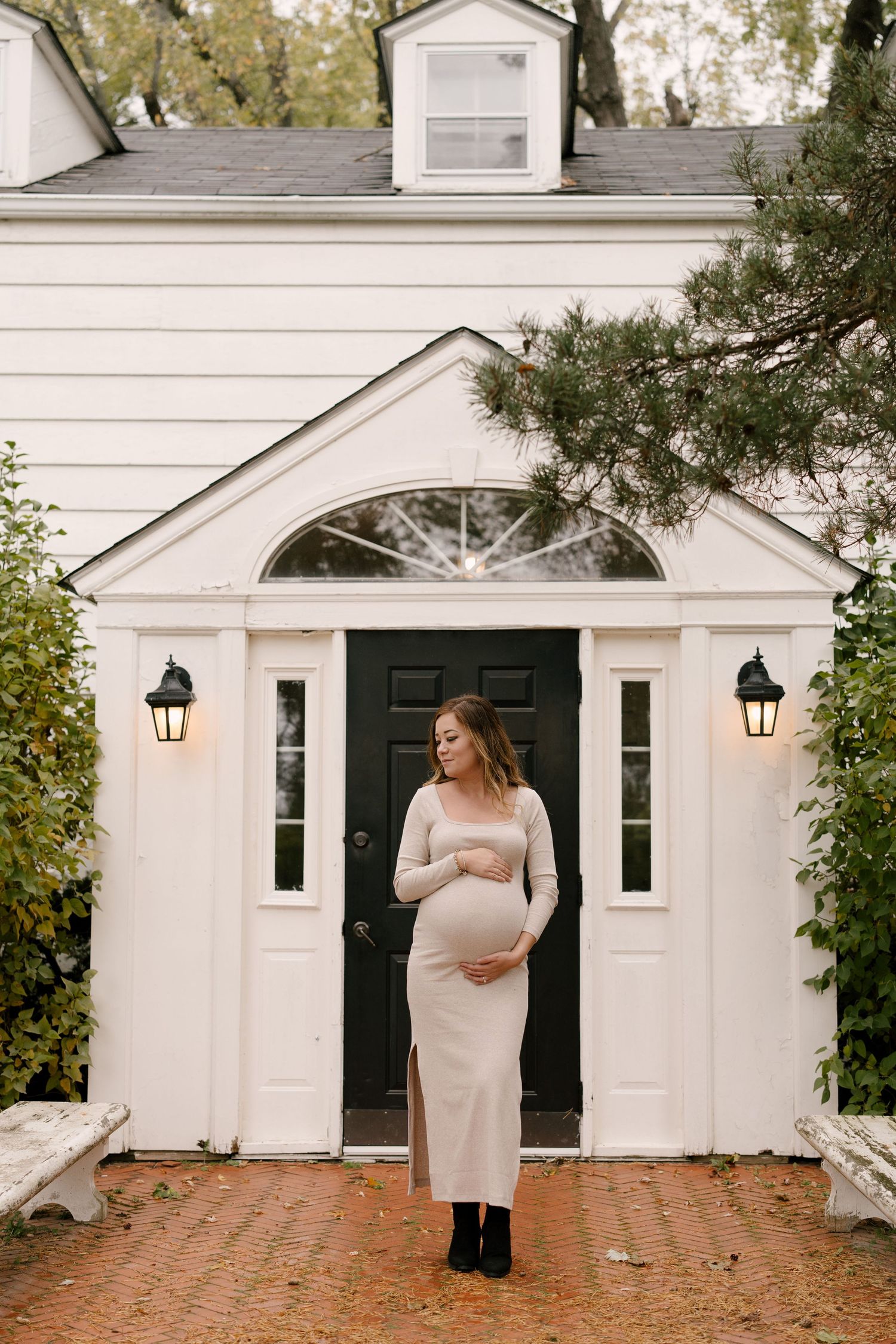 Pregnant woman in cream fitted dress cradling baby bump before elegant white colonial home.