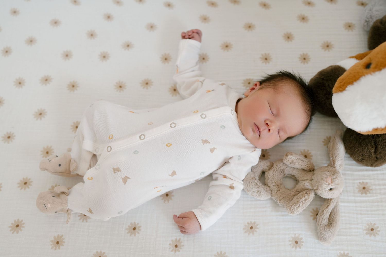 Sleeping newborn baby in white floral footie pajamas lying on daisy print crib sheet with stuffed animals.