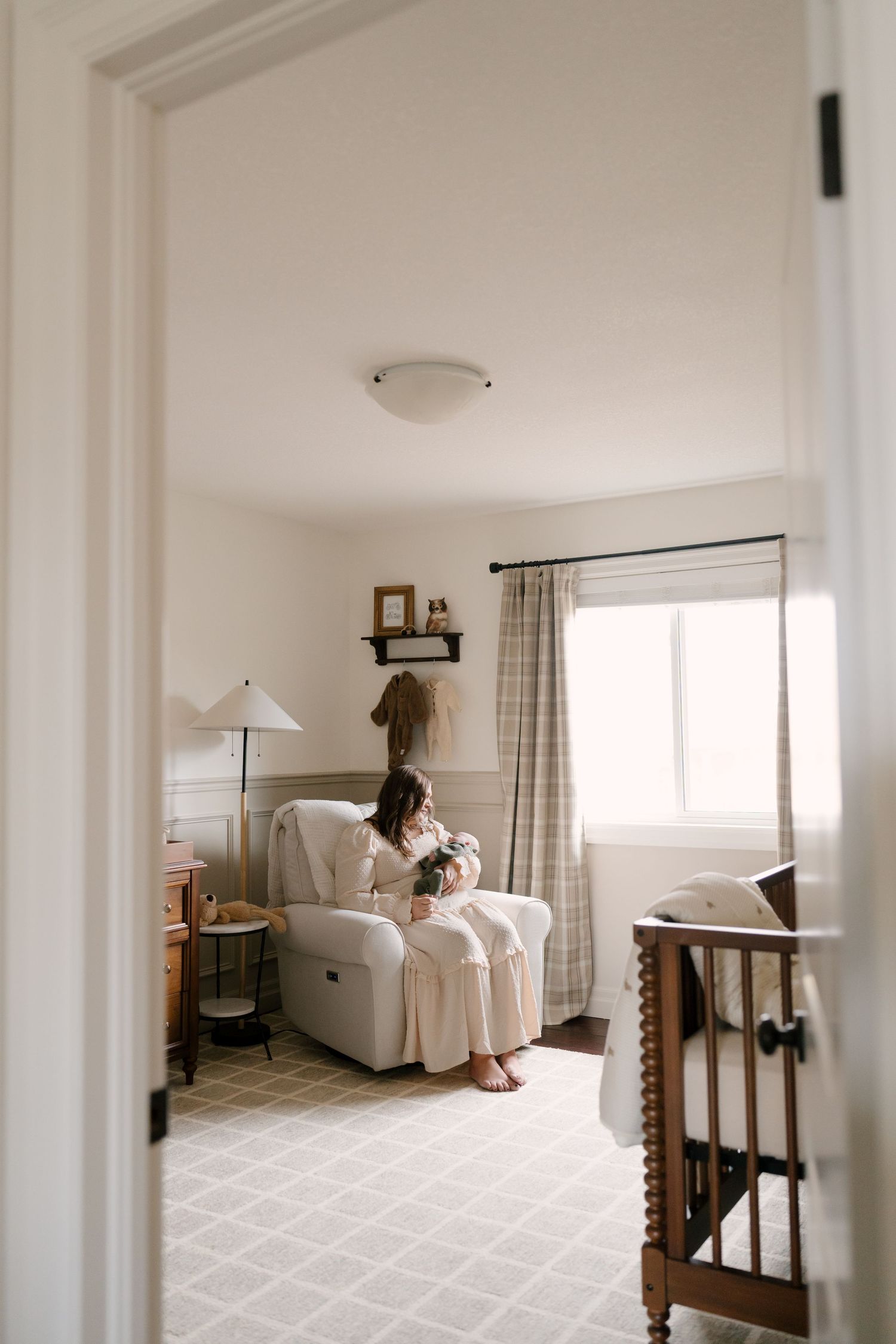 Mother sits in a glider chair nursing her newborn in a soft, neutral-toned nursery room.