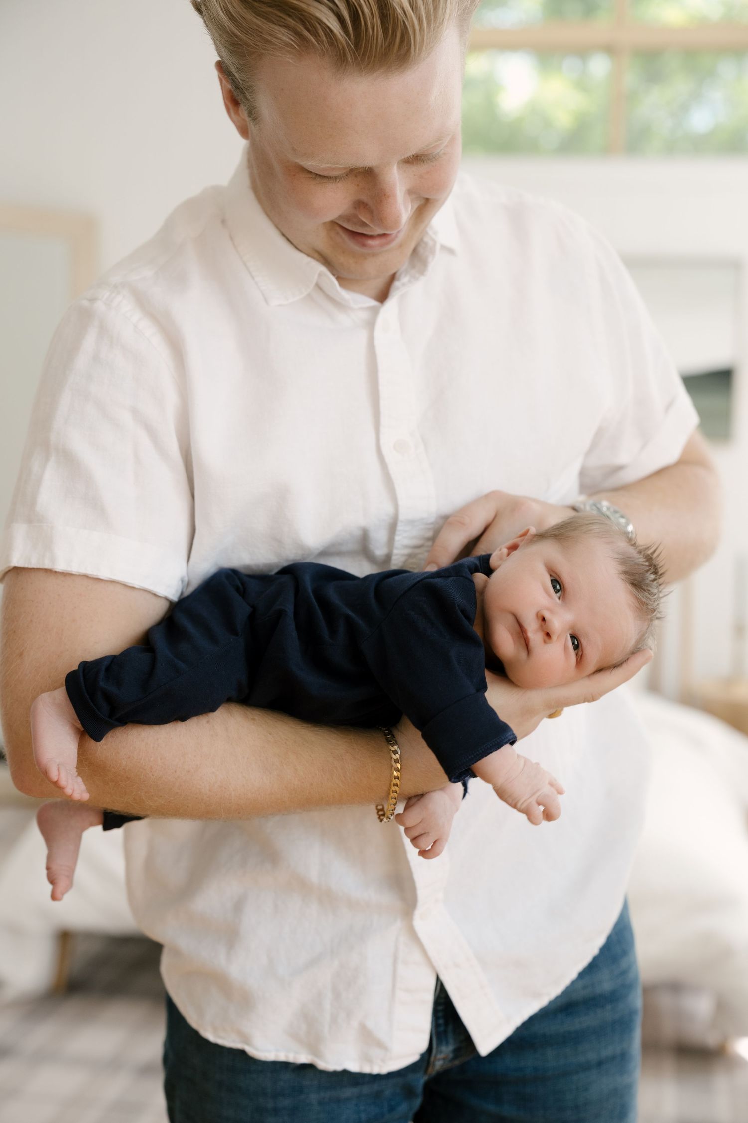 A smiling father in a white shirt holds his newborn baby dressed in navy in a bright, cozy room.