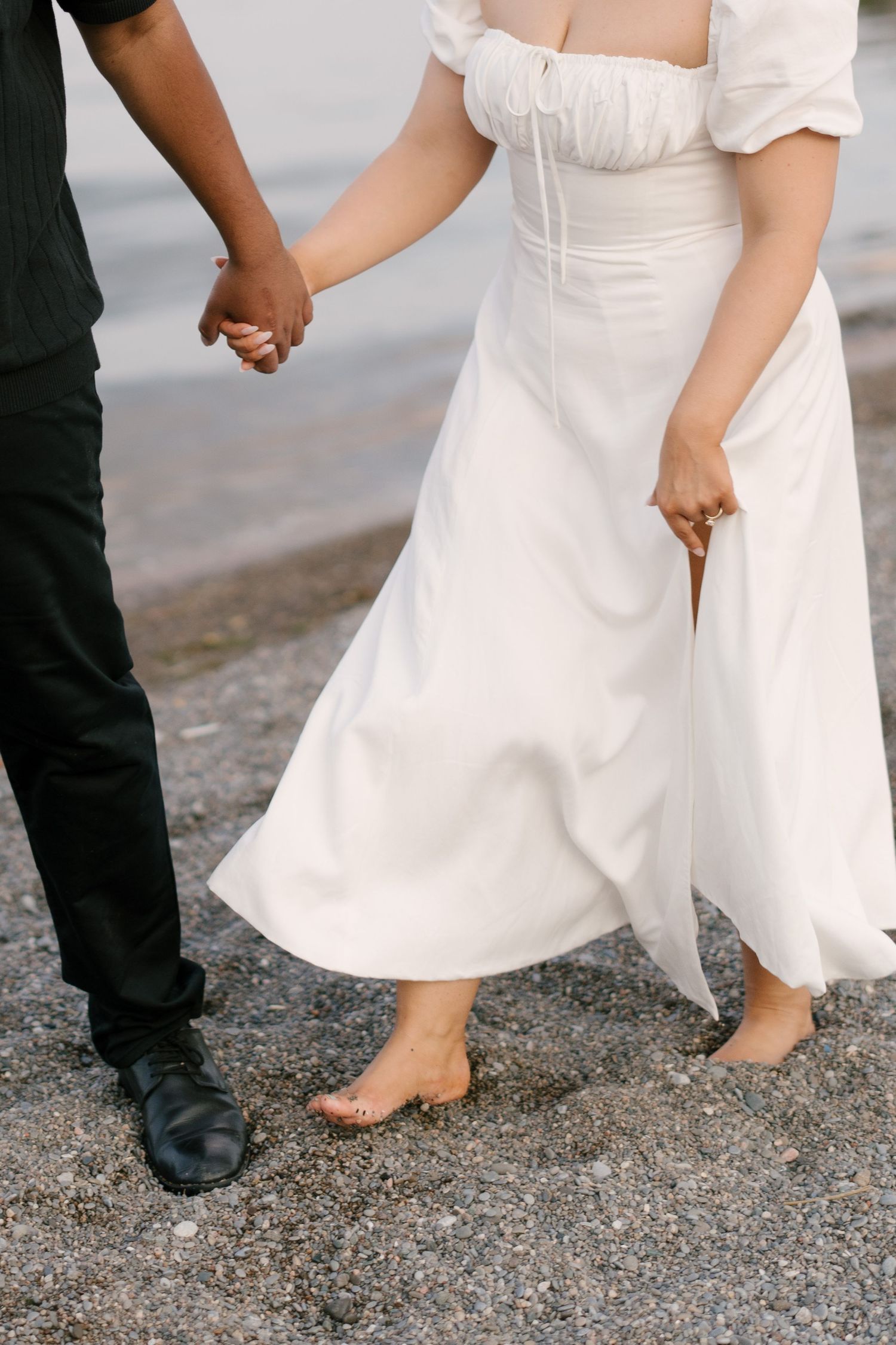 A couple walks hand in hand along a sandy beach shoreline, the woman wearing a flowing white dress.