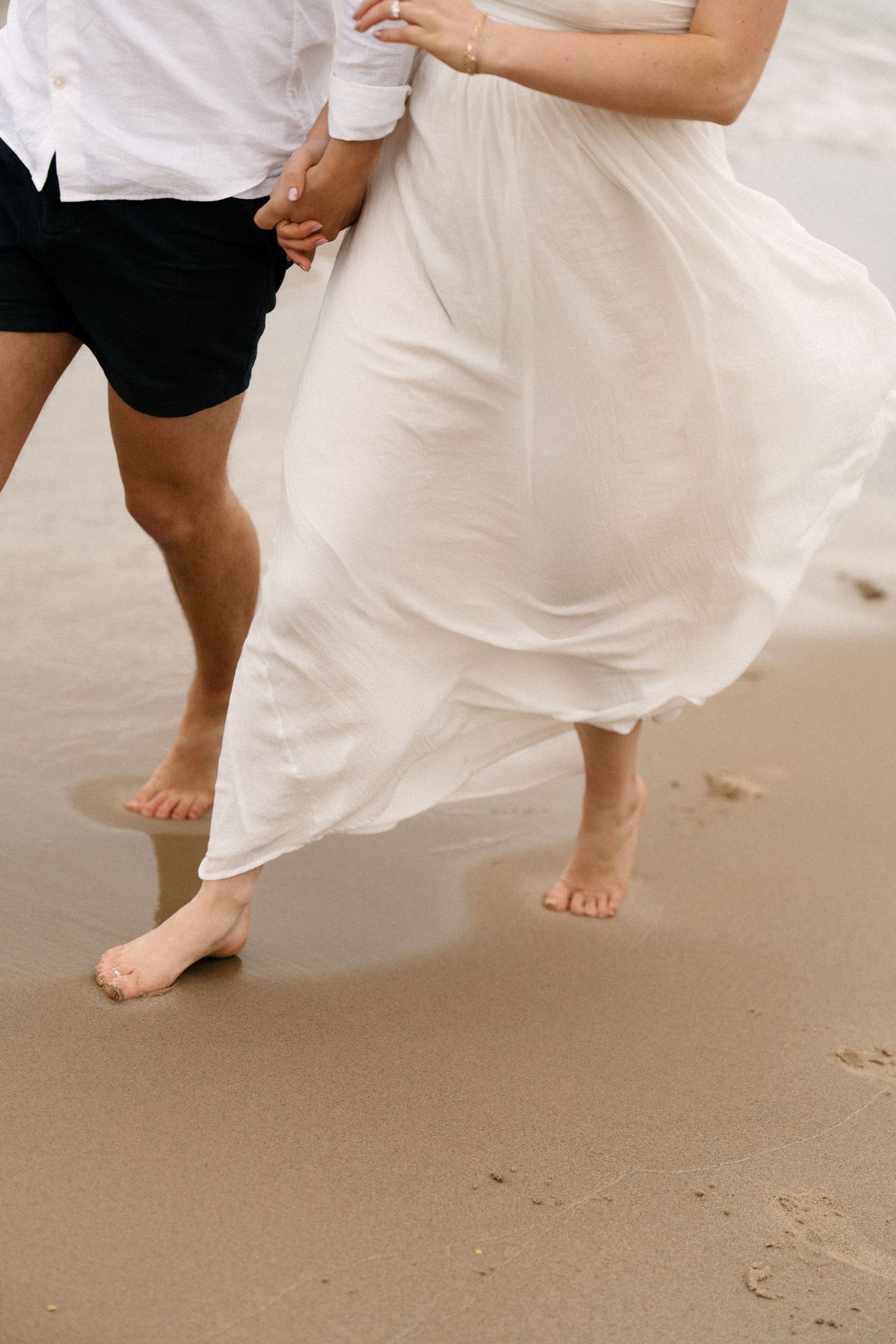 Close-up of barefoot couple walking on wet sand, white flowing dress billowing in the breeze near ocean waves.