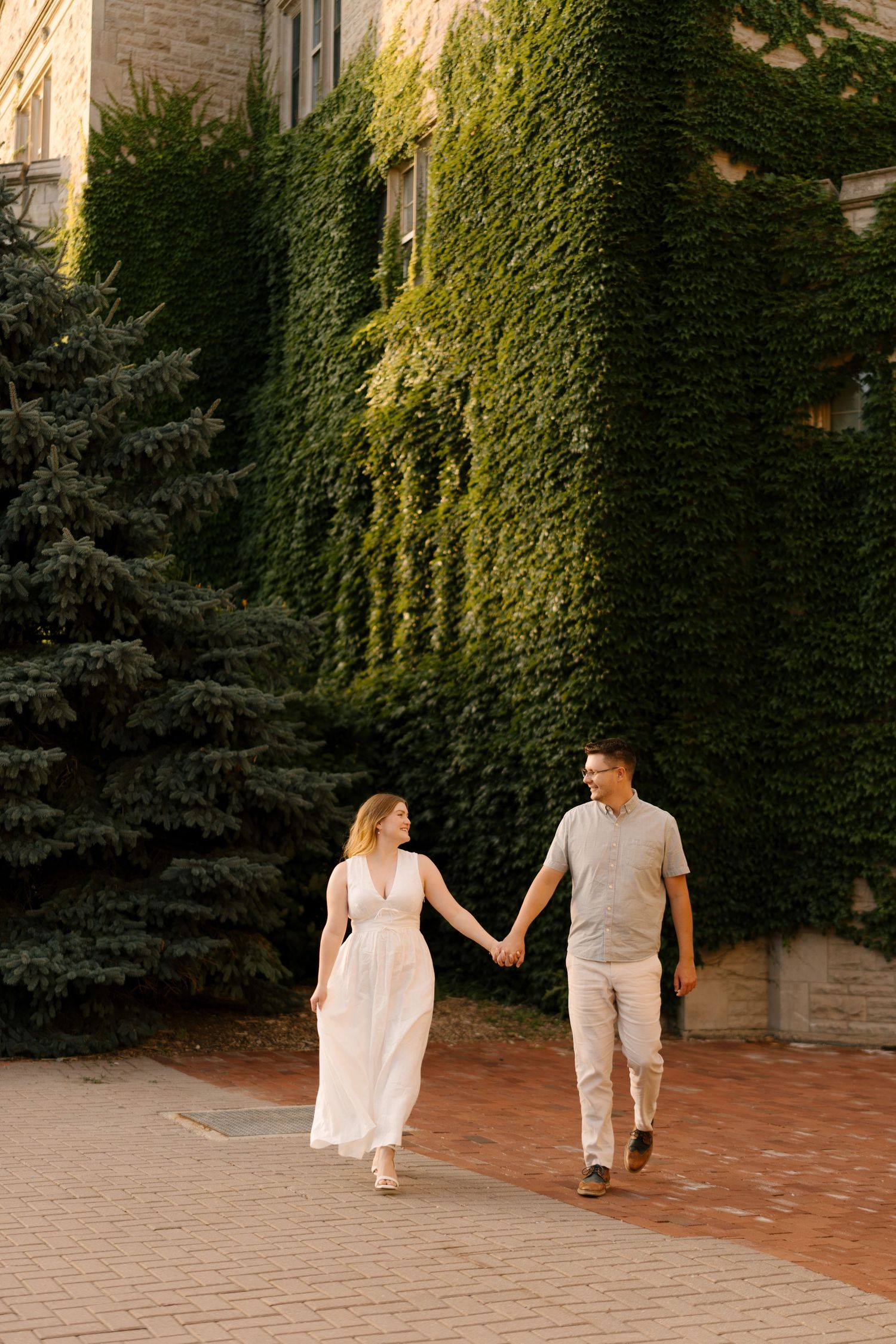 Couple walking hand-in-hand on brick path past ivy-covered stone building in golden afternoon light.