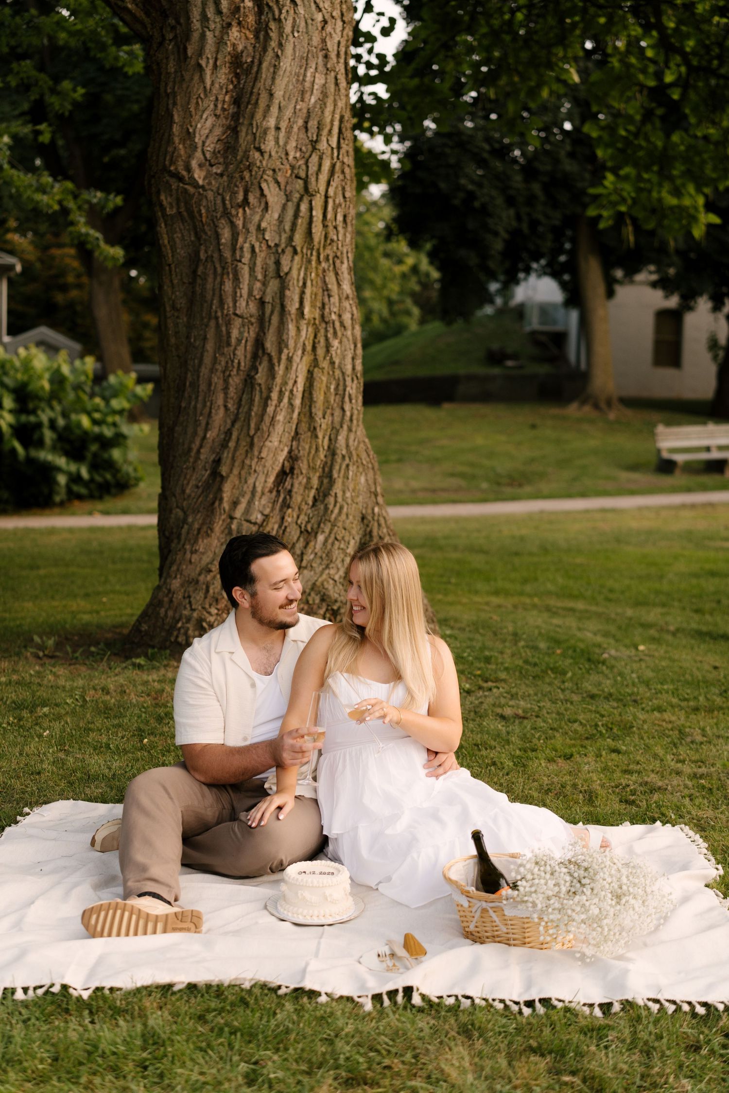 Smiling couple sitting on a white blanket under a large tree with champagne and a white cake.